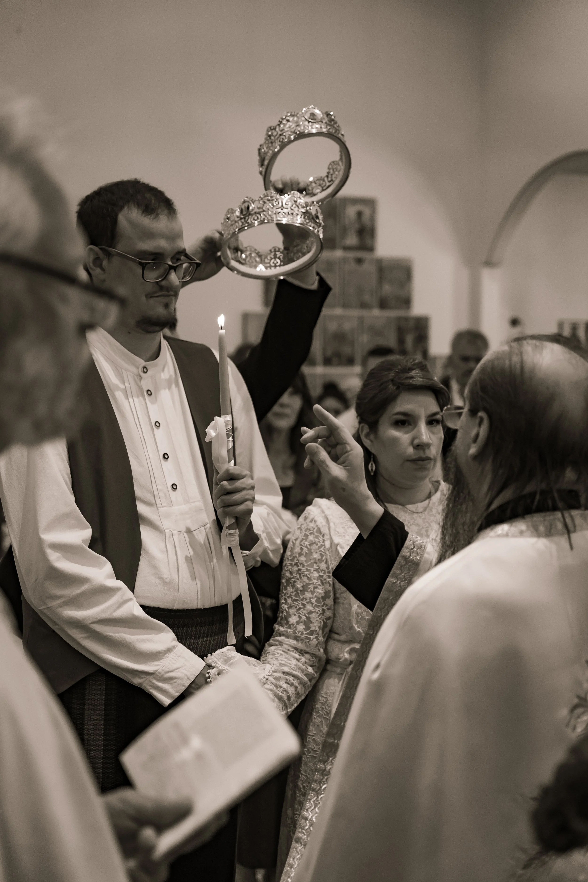 A man holding two ornate crowns above his head during a ceremony, surrounded by women dressed in traditional attire, in an indoor setting with an audience in the background.