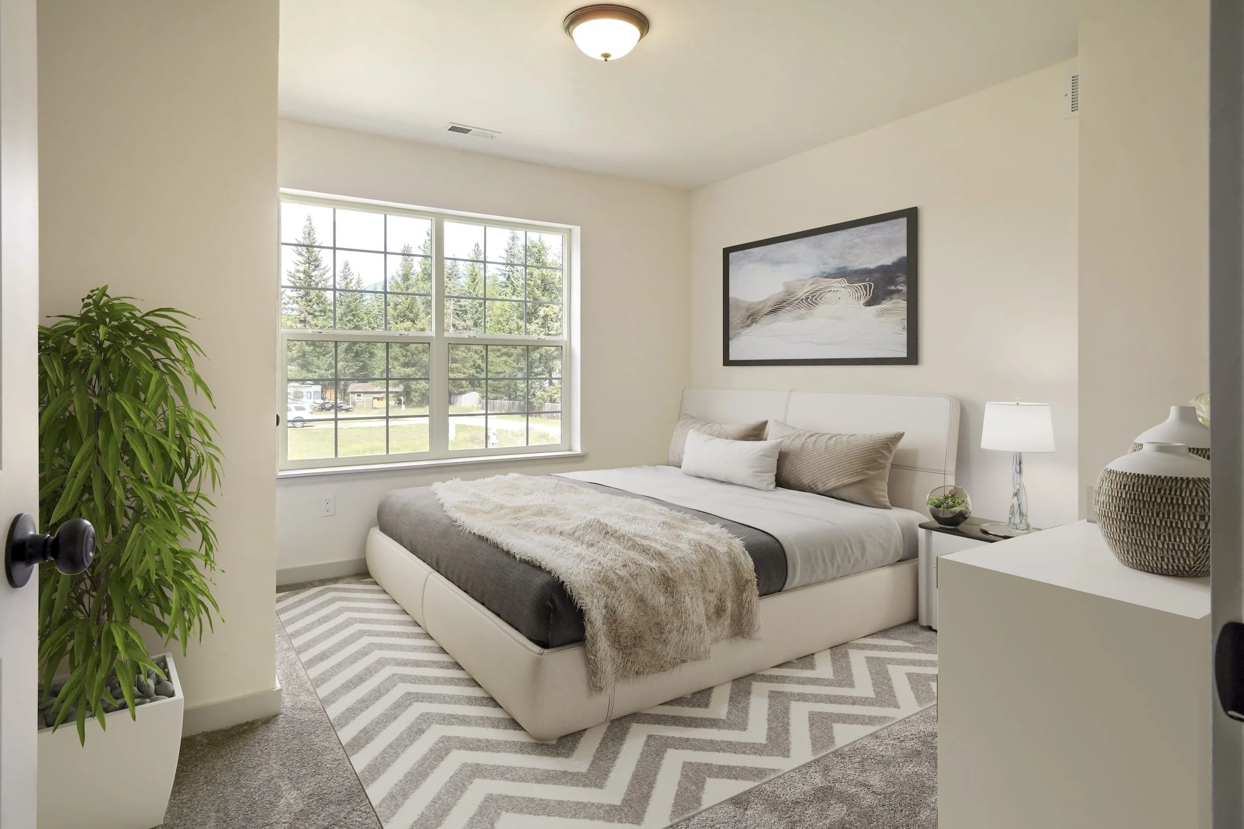 A bedroom with a large window, white walls, a bed with neutral-colored pillows and a blanket, a nightstand with a lamp, a framed artwork on the wall, a potted plant, and a patterned rug.
