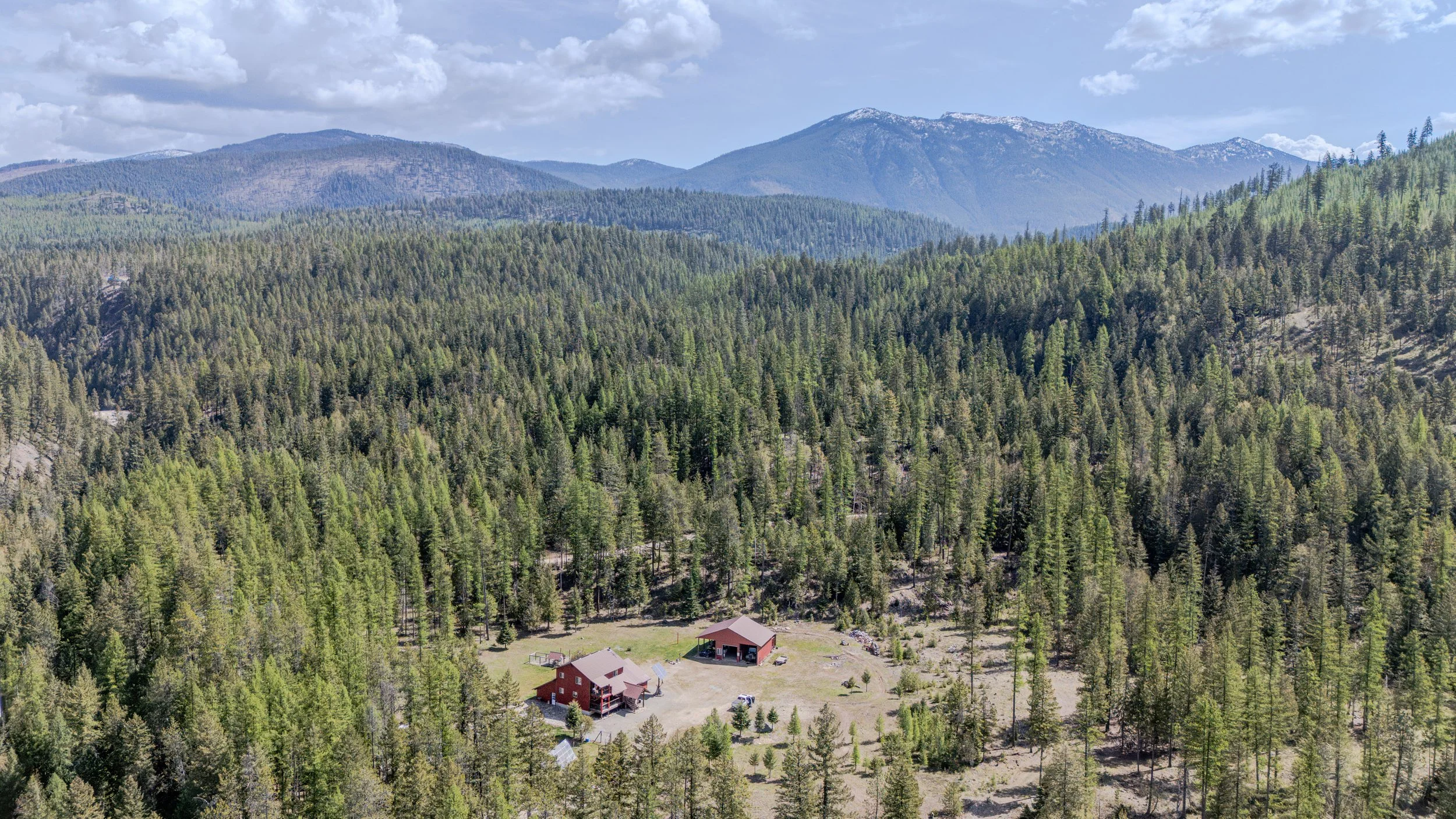 Aerial view of a rural area surrounded by dense green trees and forested mountains in the distance, with a few houses visible in the foreground.
