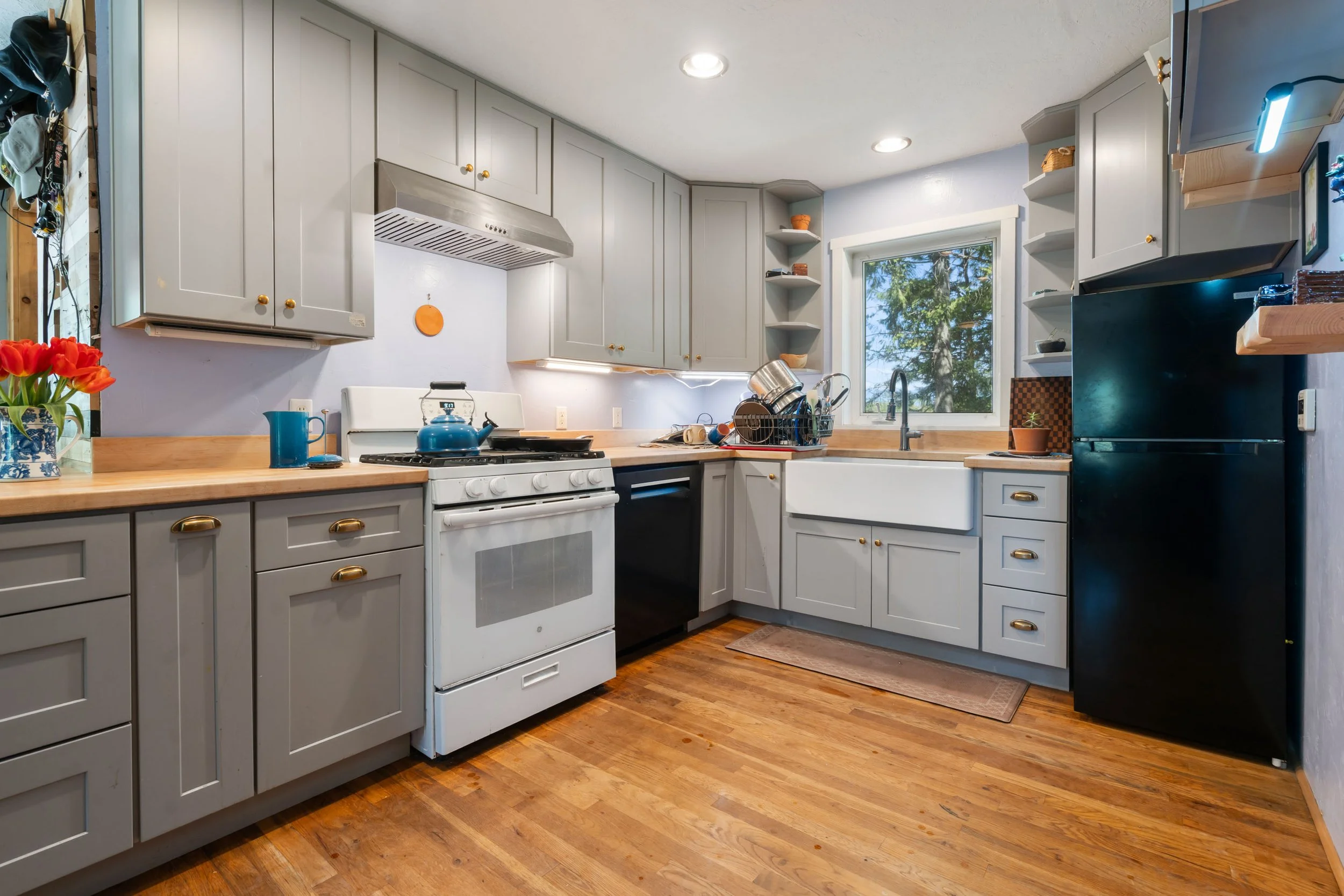 A kitchen with light gray cabinets, a white stove, a wooden countertop, and hardwood flooring. There is a window above the sink showing trees outside, and various kitchen items including a blue kettle and dish rack.