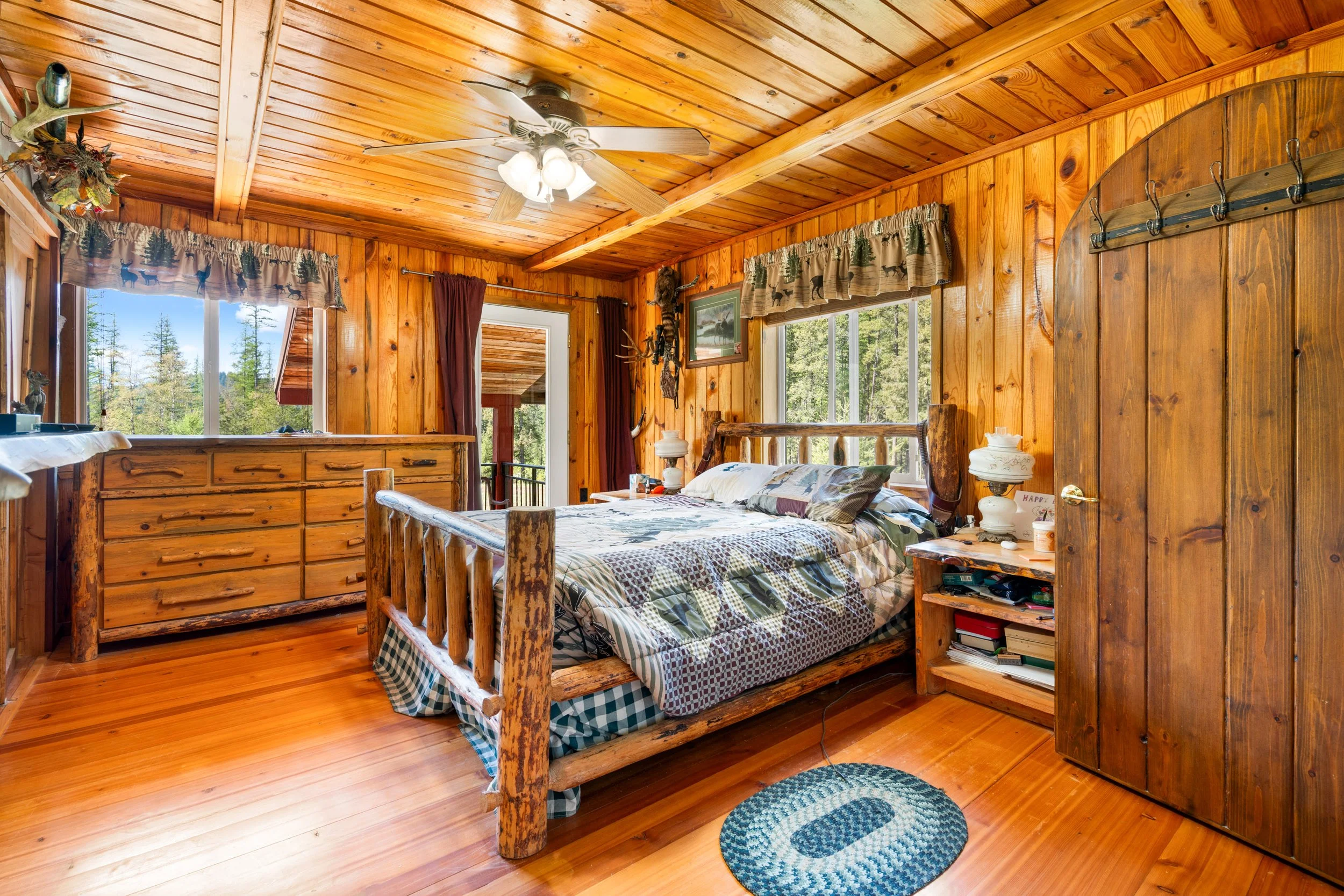 Cozy rustic bedroom with wood-paneled walls and ceiling, featuring a log bed with plaid bedding, windows with wilderness-themed curtains, and outdoor scenery visible outside.