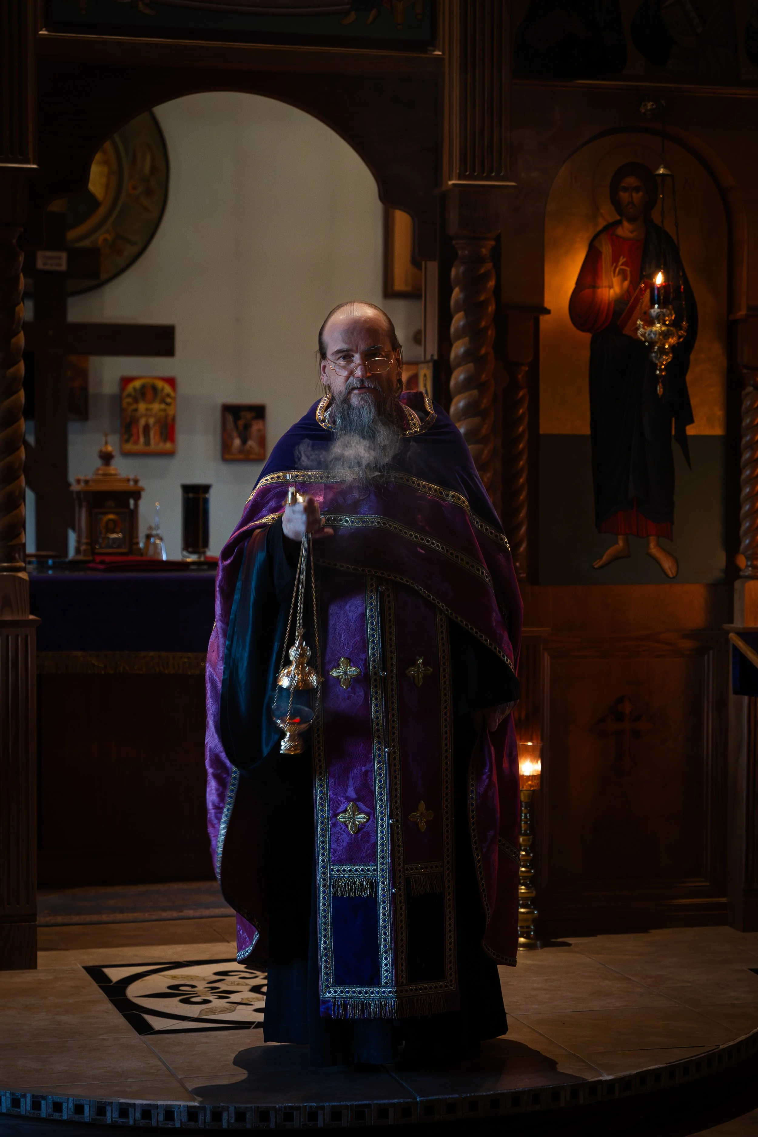 An Orthodox priest in a dark purple and gold vestment stands inside a church, holding a censer. Behind him, an icon of Jesus Christ is mounted on a wooden wall with ornate columns, and a candle holder with lit candles is visible.