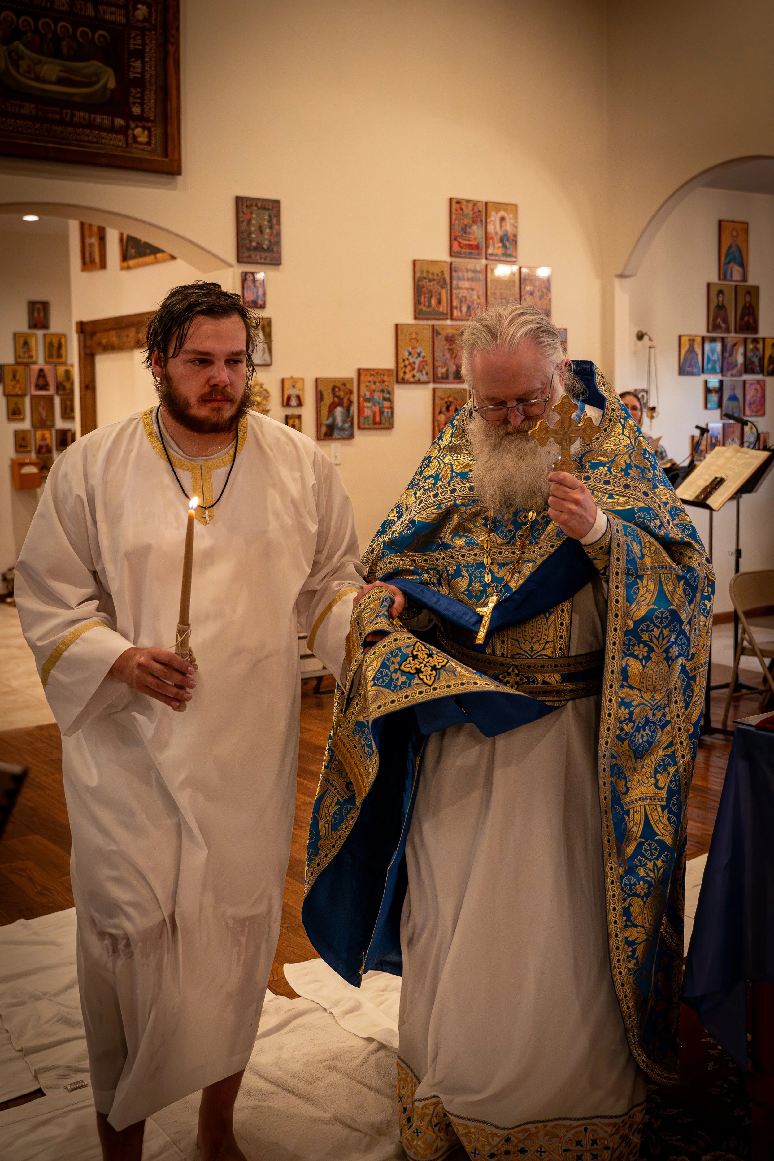 A religious ceremony with two men in traditional religious robes inside a church. One man is holding a candlestick with a lit candle, and the clergy is holding a cross and wearing ornate blue and gold robes. The background has numerous religious icon