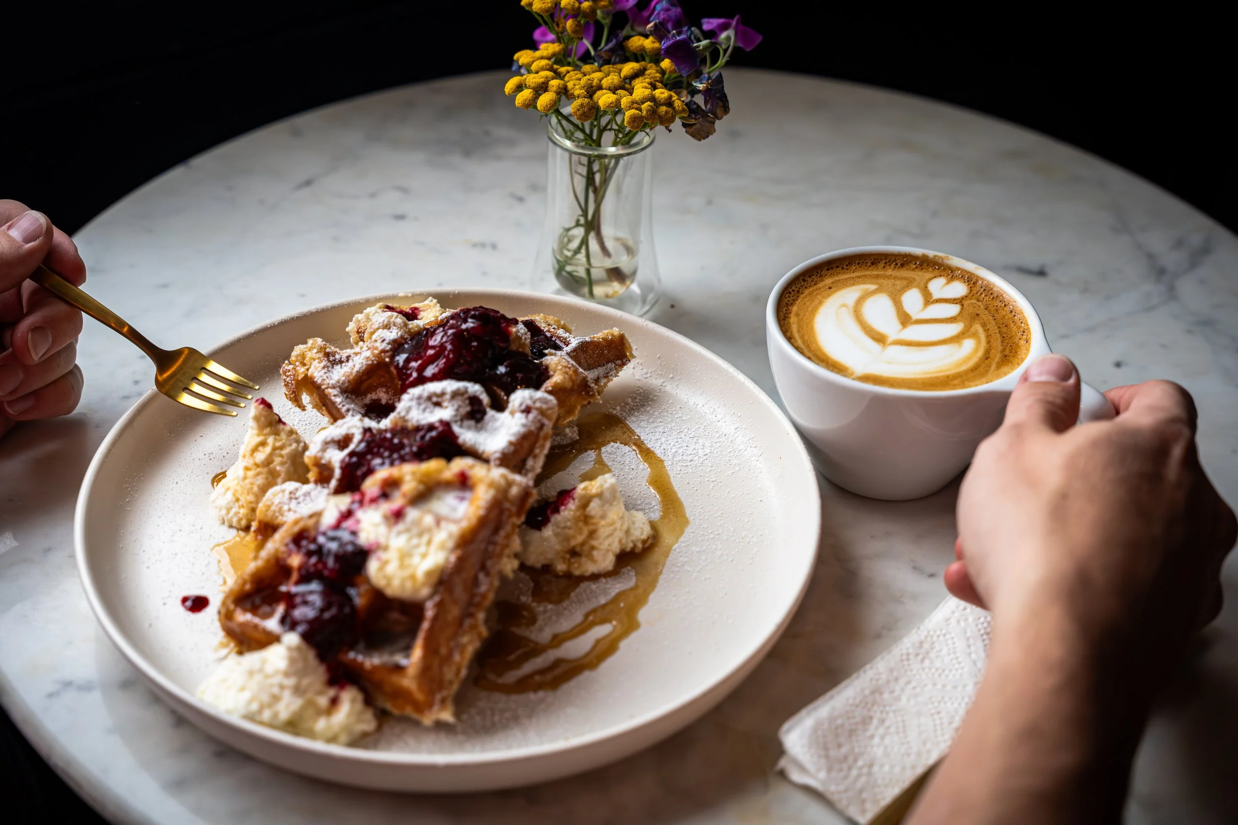 A person holding a fork over a plate with a slice of cherry pie topped with powdered sugar, alongside a scoop of vanilla ice cream, on a marble table. In the background, a cup of latte with latte art and a small vase with purple and yellow flowers.