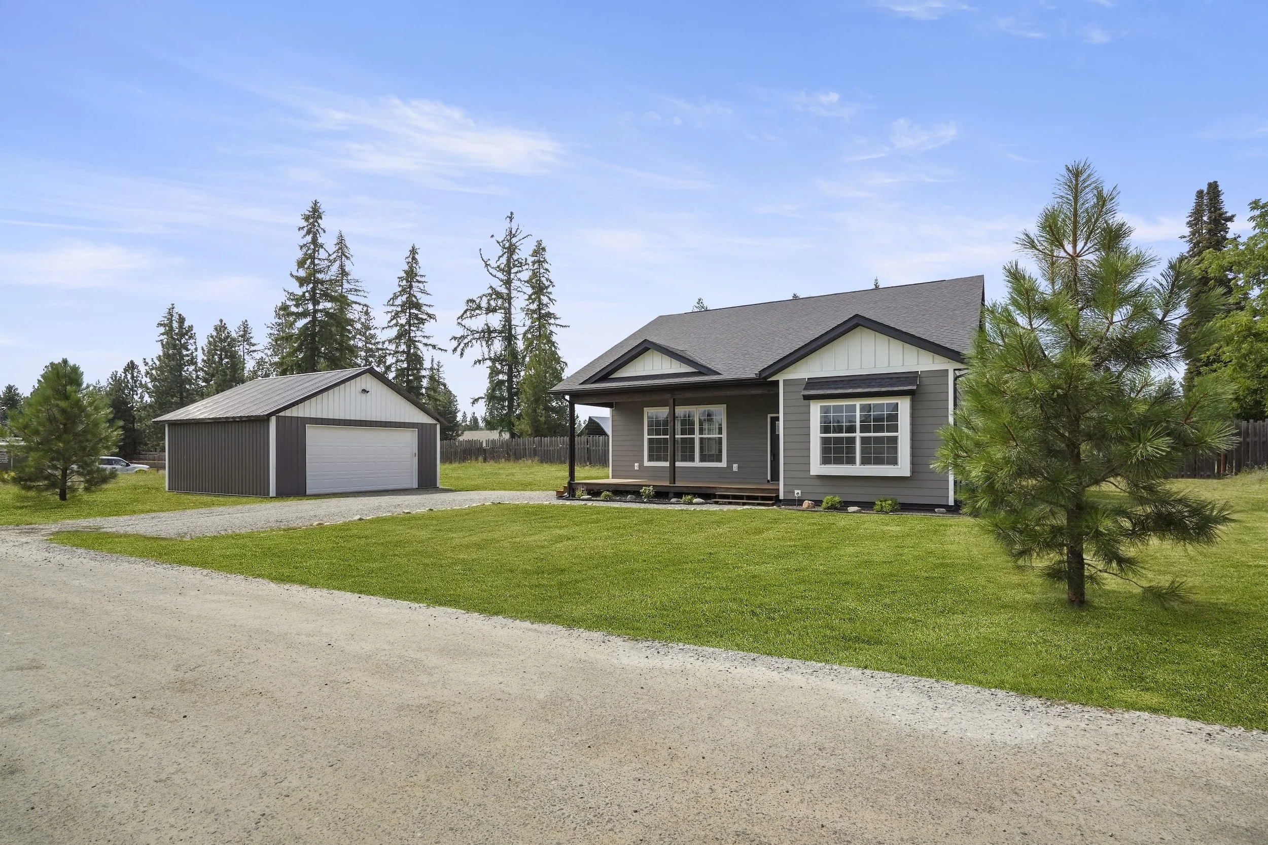 A photo of a house with a garage in a rural setting, surrounded by tall trees, with a gravel driveway and a grassy yard under a partly cloudy blue sky.