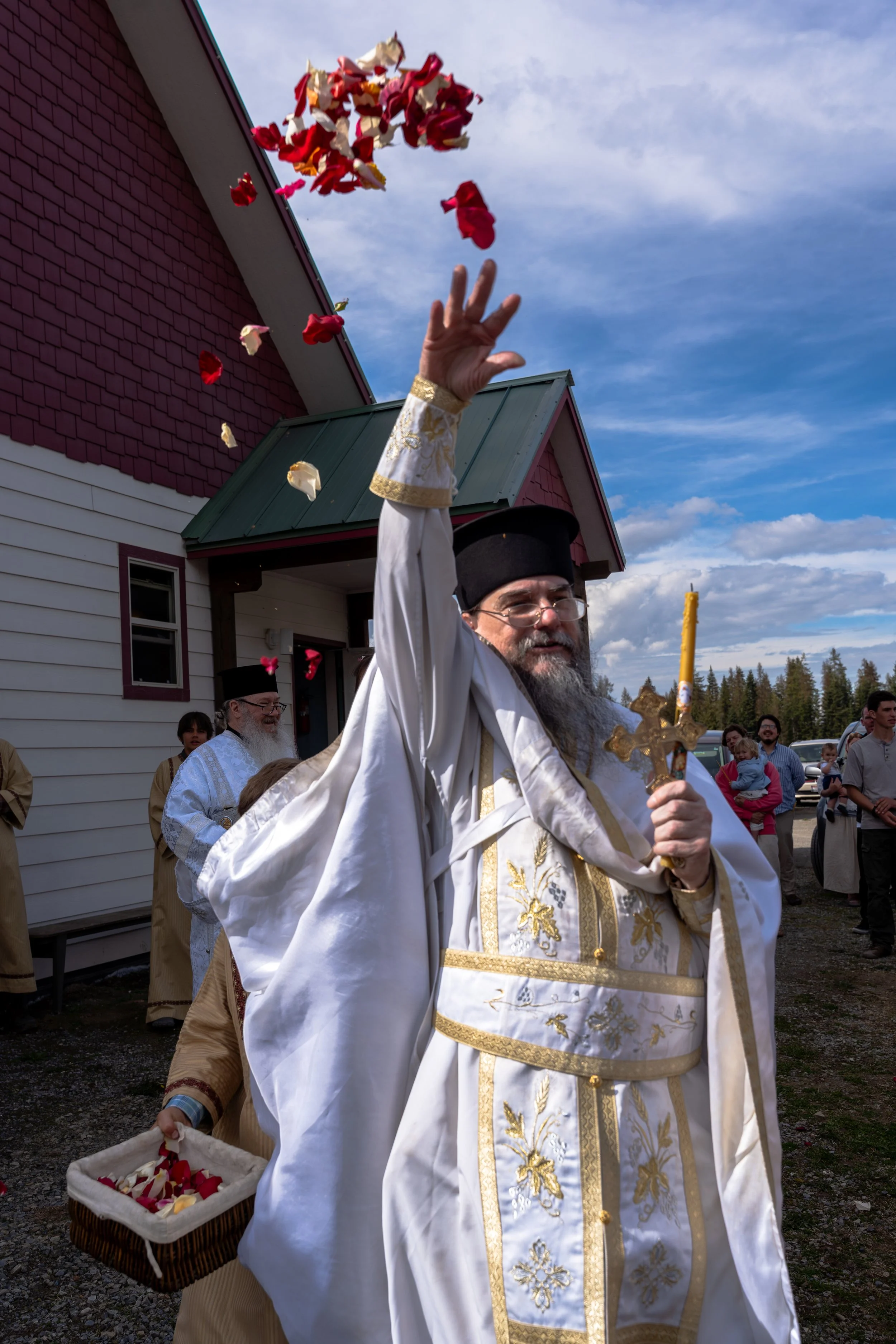A man dressed as a priest or religious figure wearing white and gold robes, holding a cross and a candle, participating in a religious outdoor procession with flower petals falling from above, on a sunny day with a blue sky and a small crowd in the b