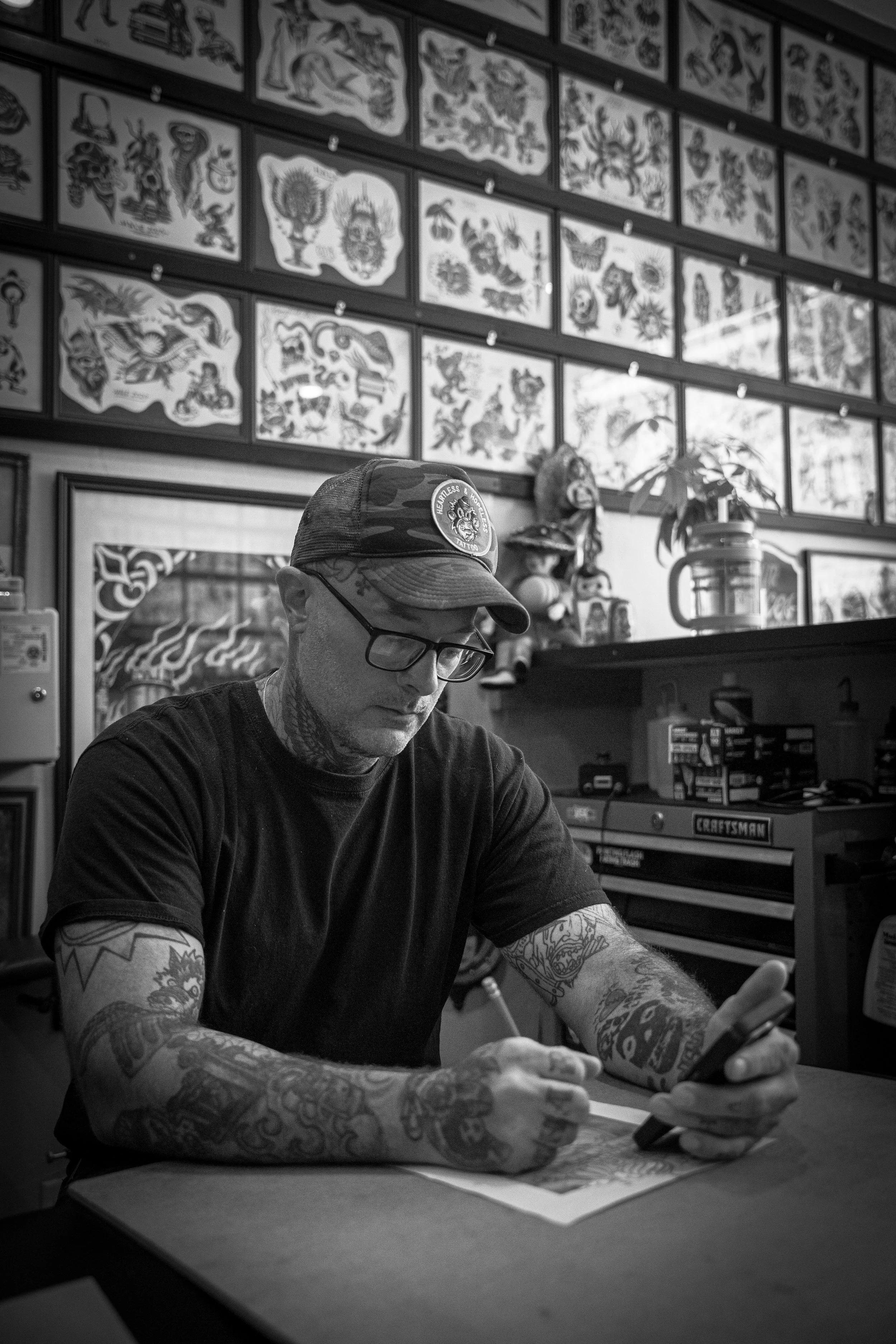 A person wearing a cap and glasses, with tattoos on their arms, is sitting at a table using a smartphone. The background shows a wall decorated with framed tattoo art and a cabinet with tools.