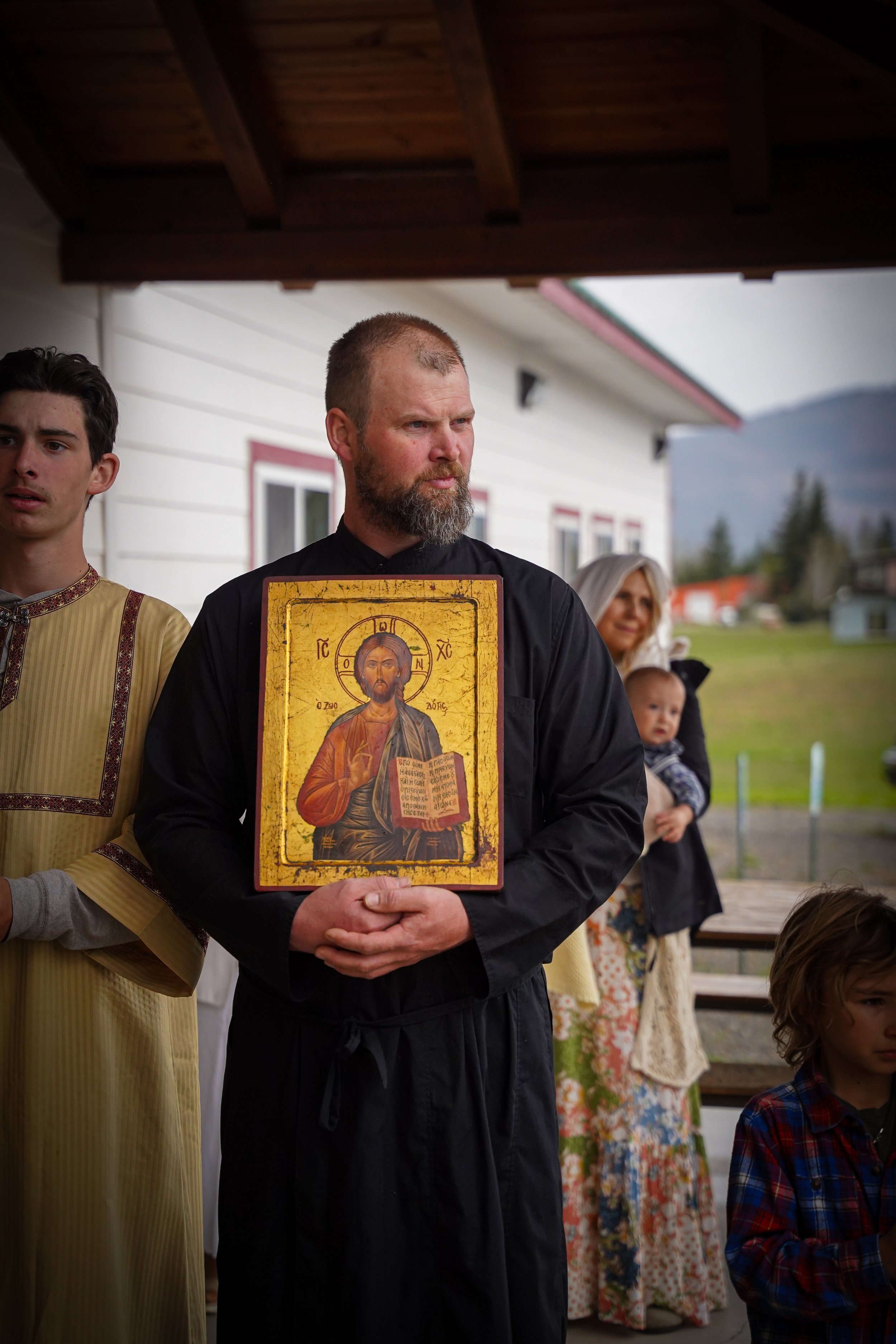 A man with a beard holding an icon of Jesus Christ, standing among other people outdoors under a canopy with a house and mountains in the background.