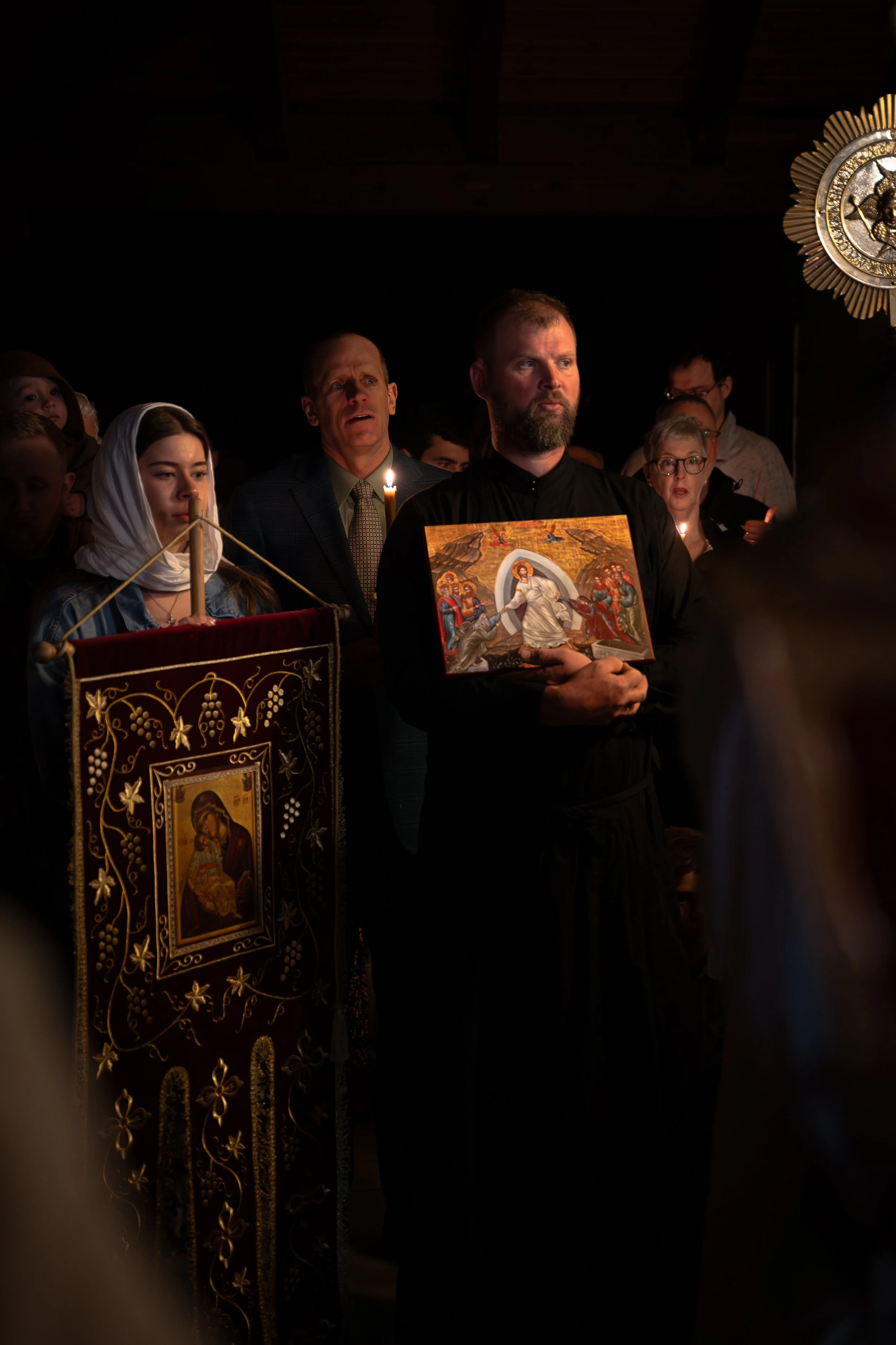 People participating in a religious ceremony, holding icons and candles, inside a dark church with religious decorations.
