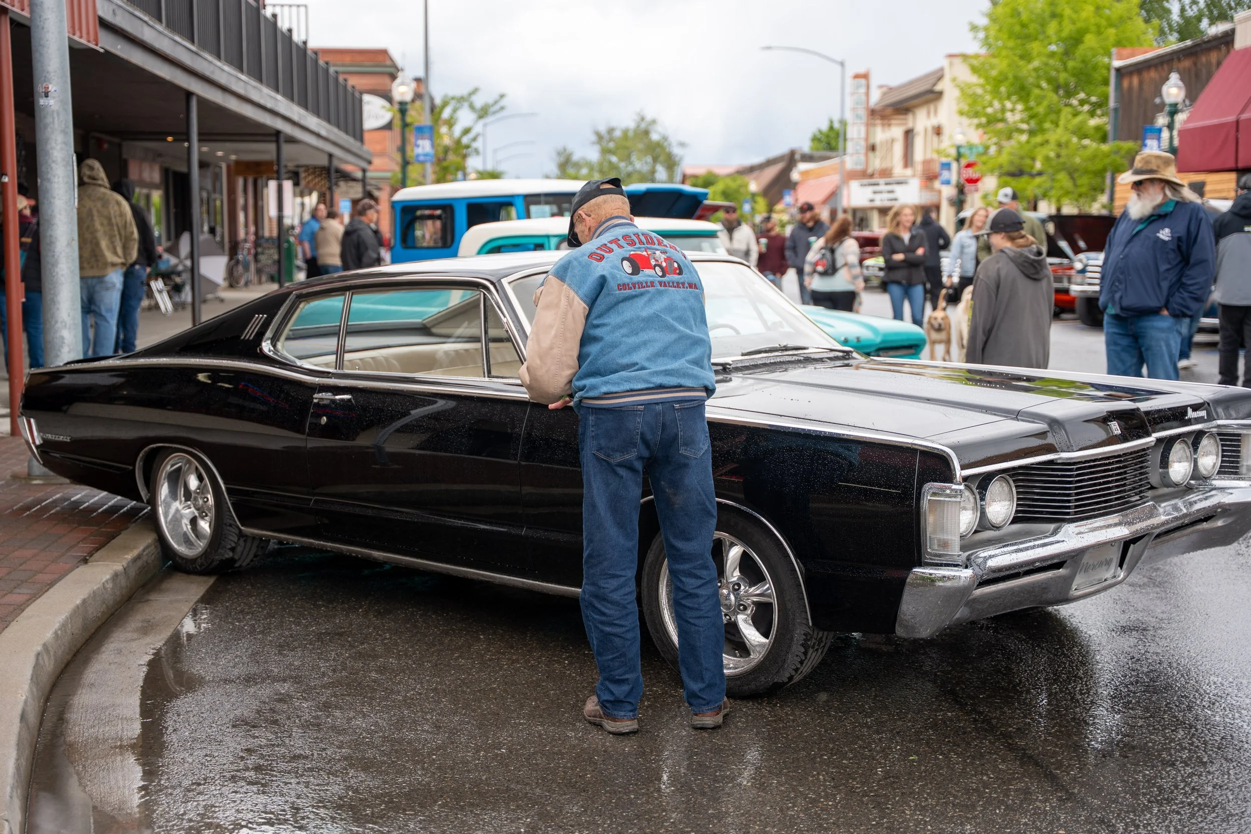 Man inspecting a vintage black car at an outdoor car show, with other classic cars and people in the background.