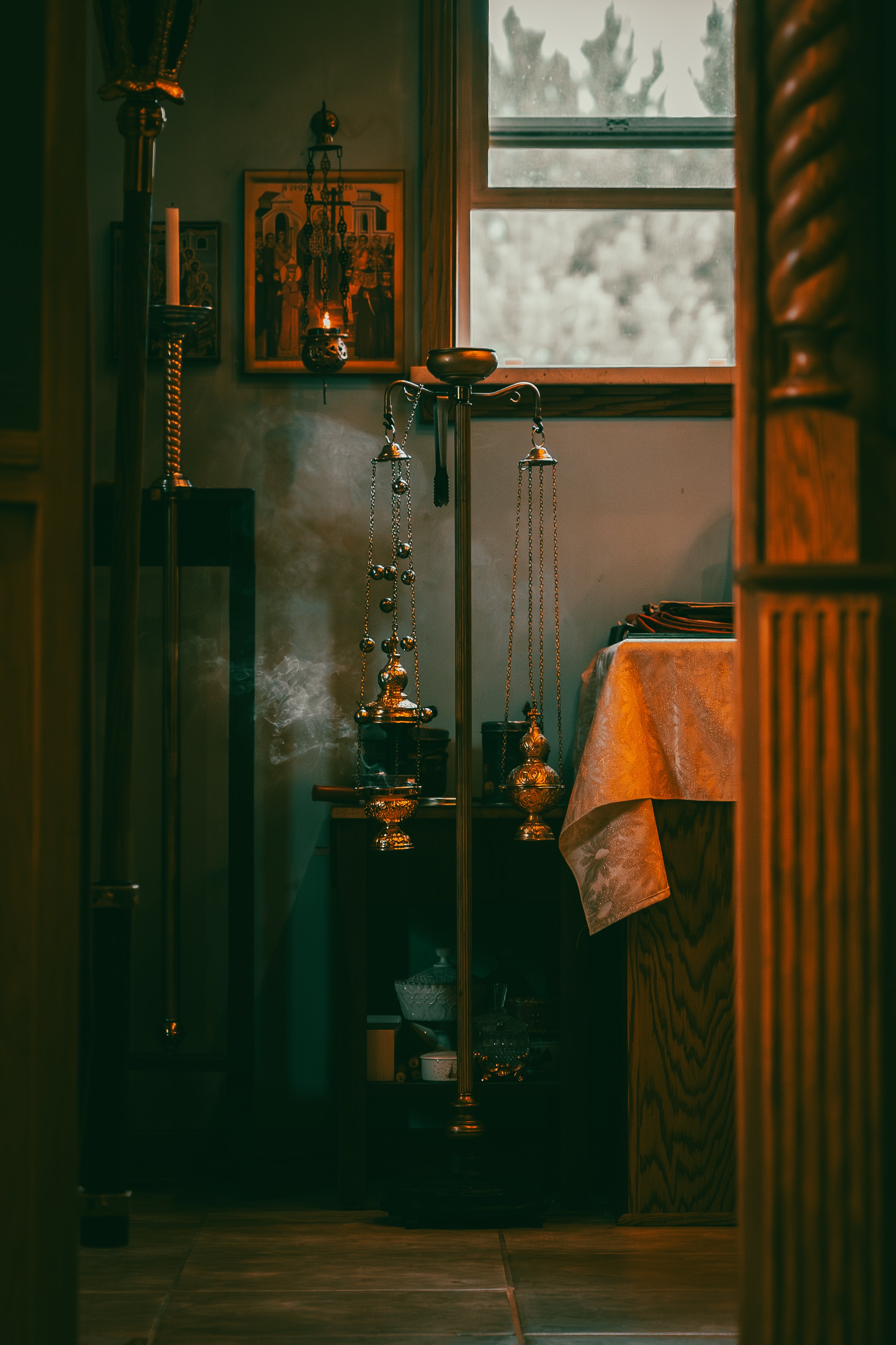 Inside a room with religious icons and gold jewelry, seen through a doorway with wooden framing and a window showing trees outside at dusk.