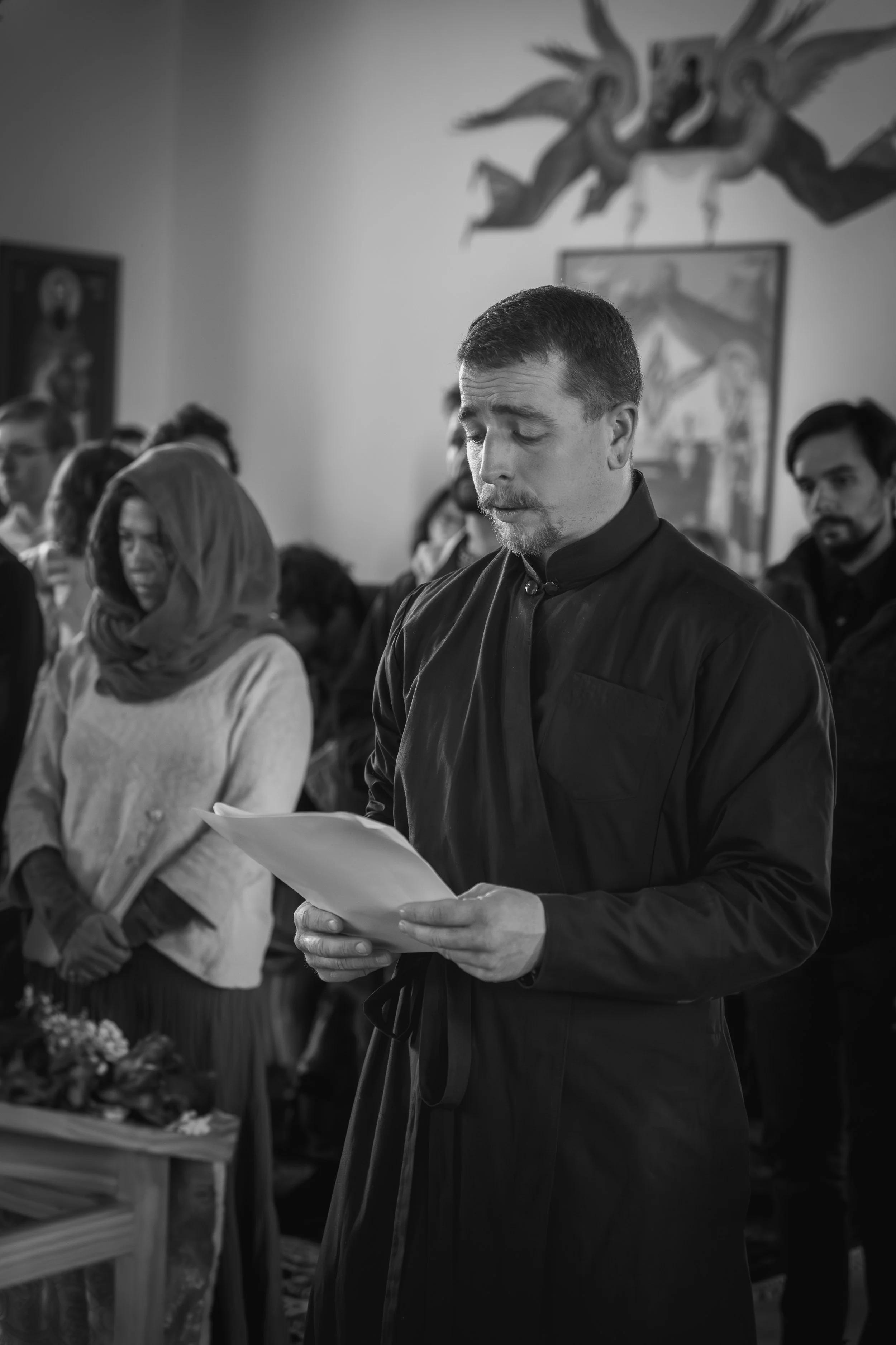 A man with short dark hair and a beard wearing a black robe, reading a paper with eyes closed or looking down, inside a room with other people, some of whom are standing with heads bowed or praying. The room has religious artwork on the walls.