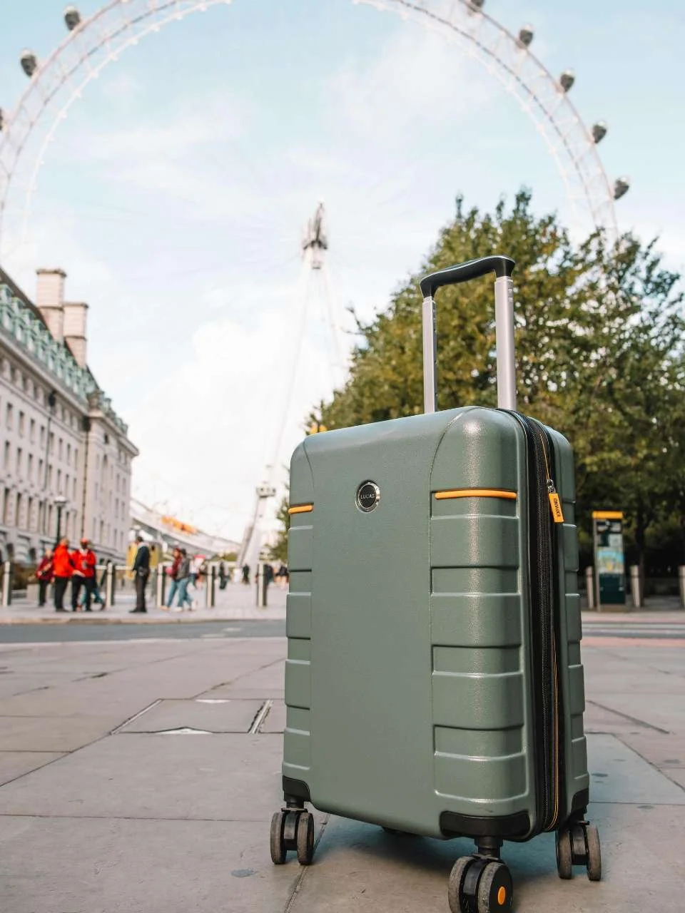 A green hard-shell suitcase with a retractable handle and four wheels on a city sidewalk, with a Ferris wheel, trees, and people in the background.