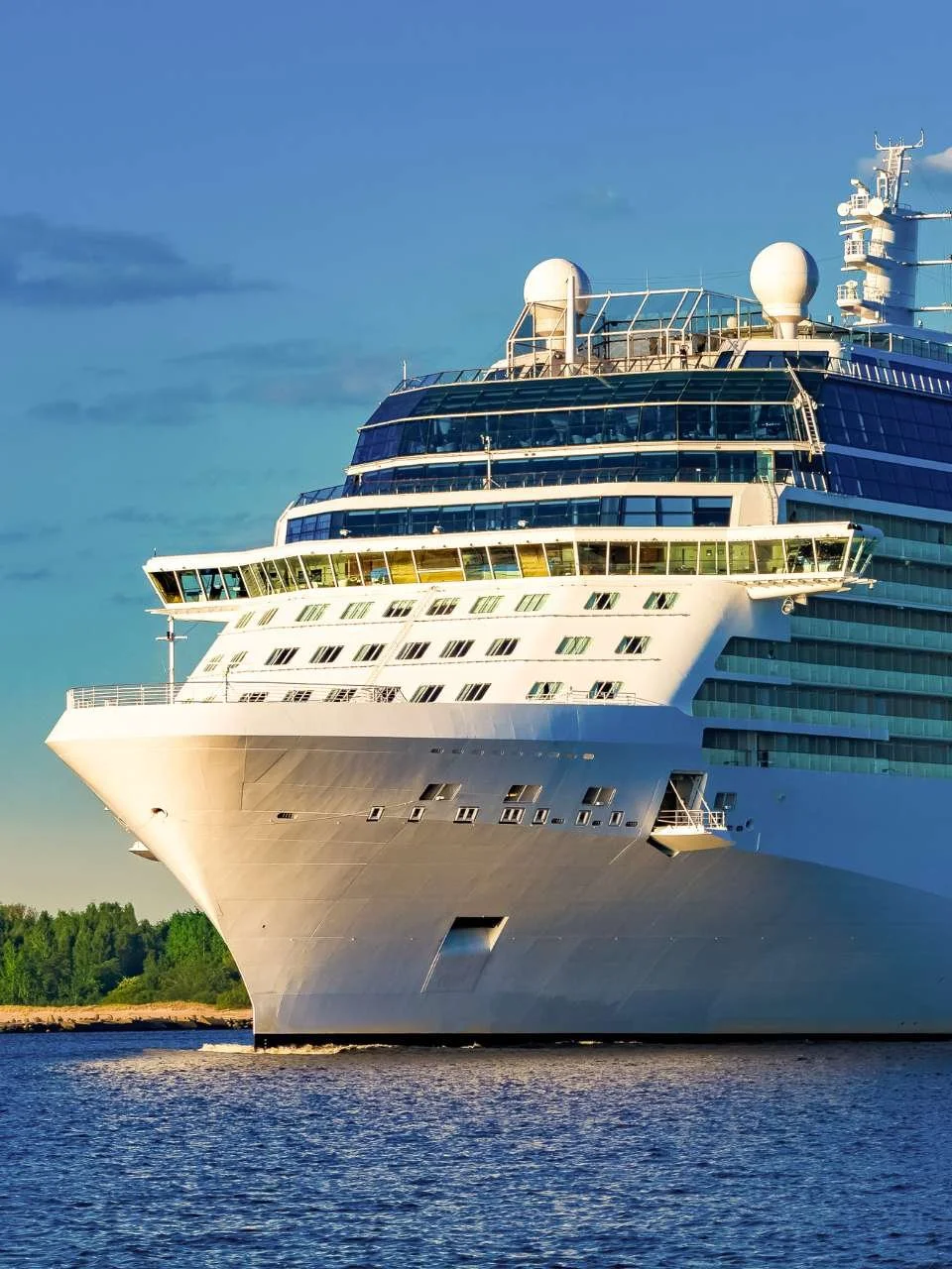 A large white cruise ship sailing near a green shoreline on a clear day.