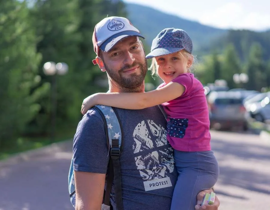 A man holding a young girl hugging him around the neck outdoors with trees and parked cars in the background.