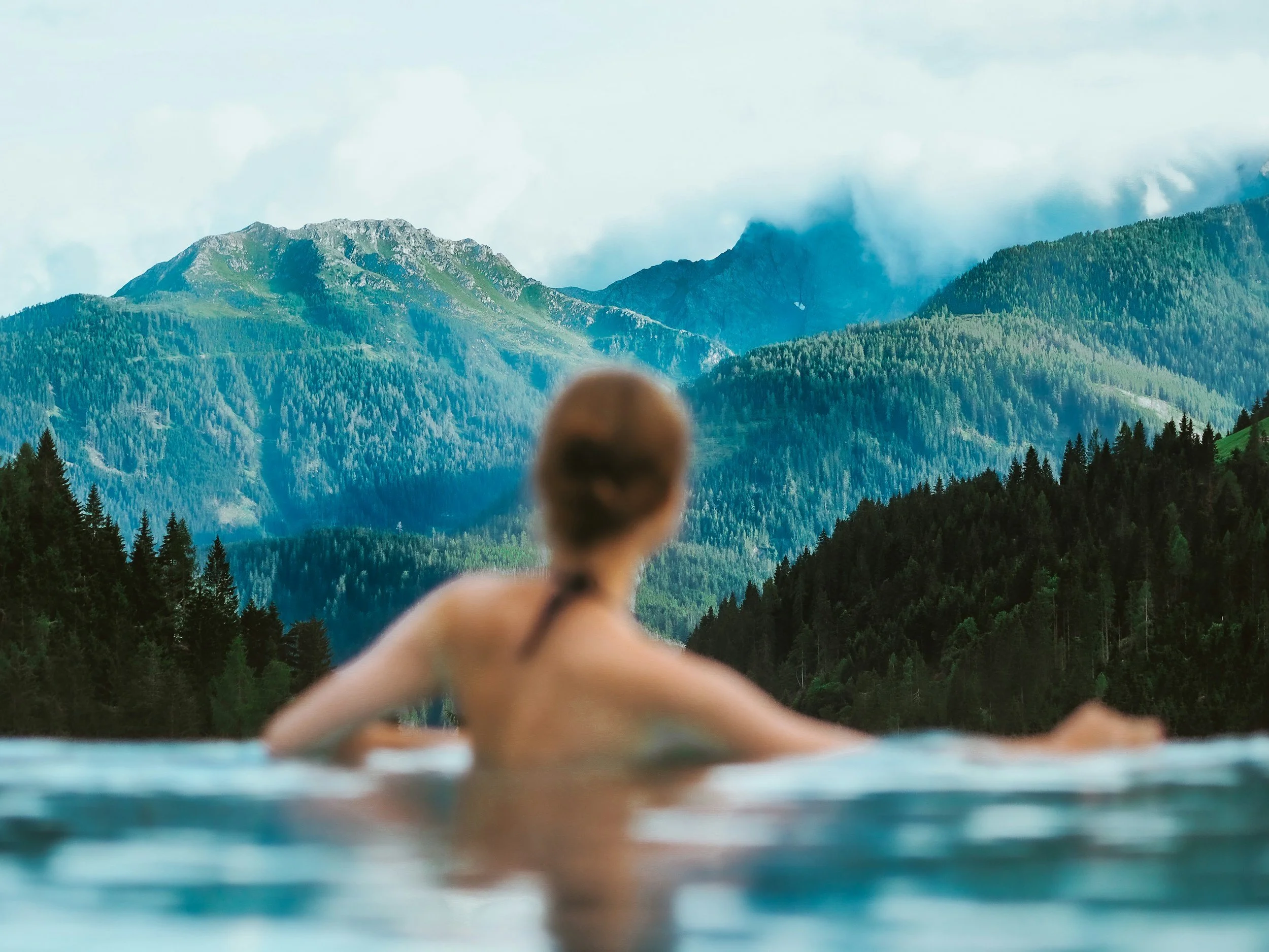Person swimming in a natural hot spring pool with scenic mountains and forest in the background.