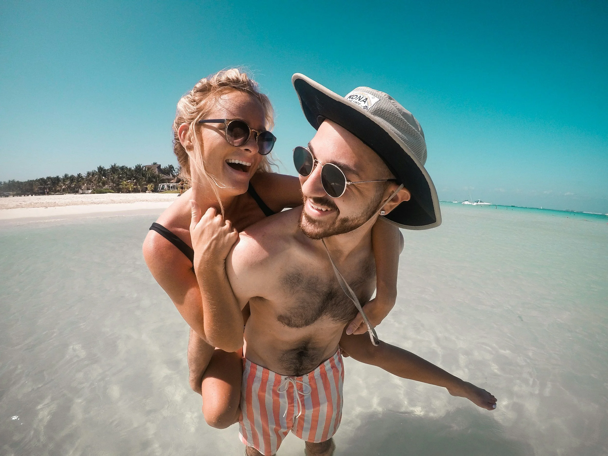 A smiling couple in swimsuits at the beach, with the woman on the man's back, both wearing sunglasses and enjoying a sunny day near the water.