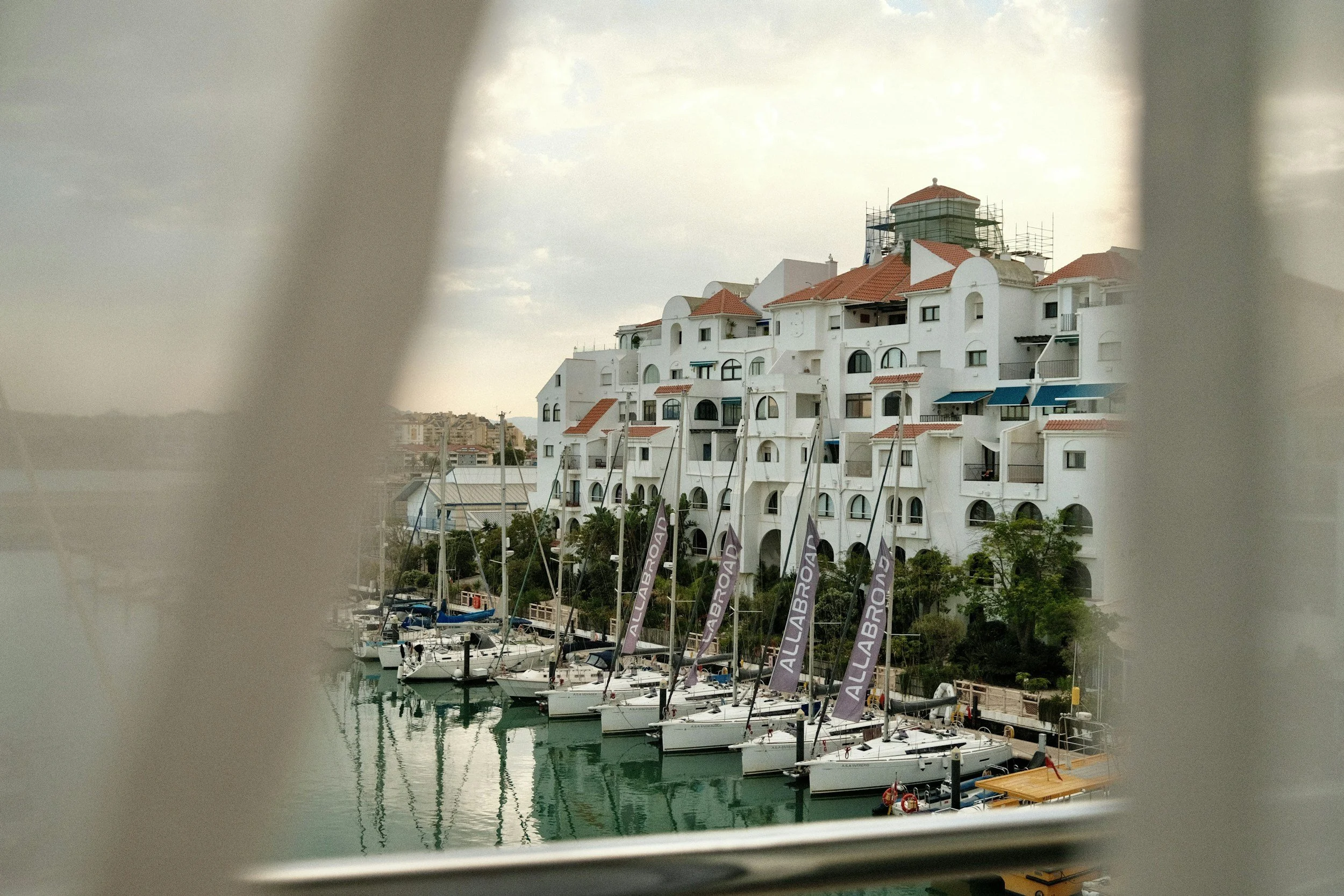 View of a marina with white sailboats docked and residential buildings with terracotta roofs in the background, framed by window blinds.