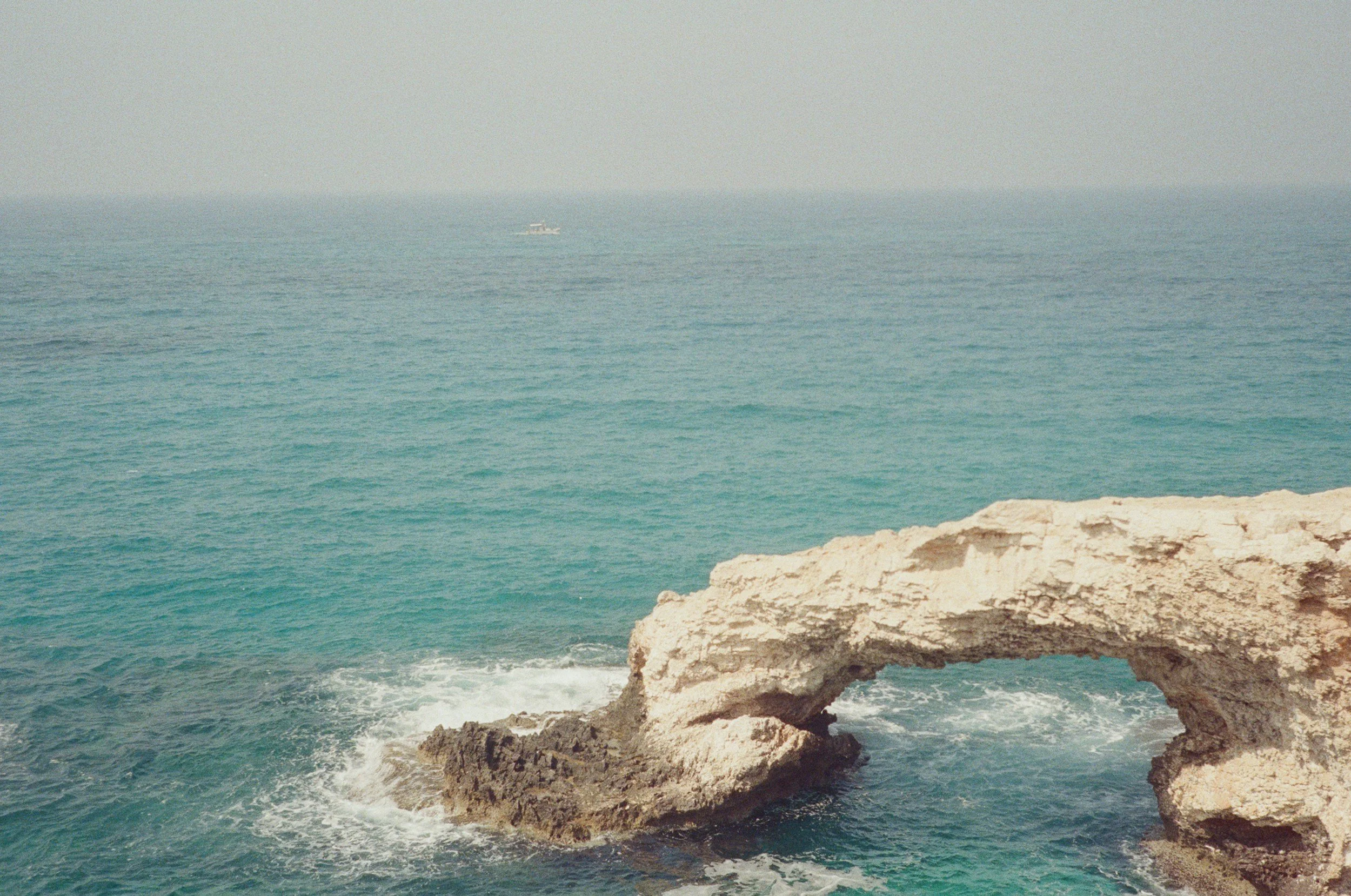 Ocean view with rocky arch formation in the water and a distant boat on the horizon.