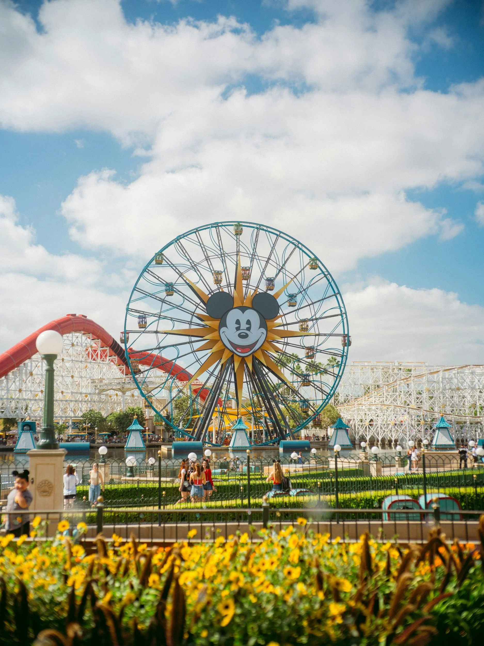 A Ferris wheel with Mickey Mouse's face at a Disney amusement park, with yellow flowers in the foreground and a partly cloudy sky overhead.
