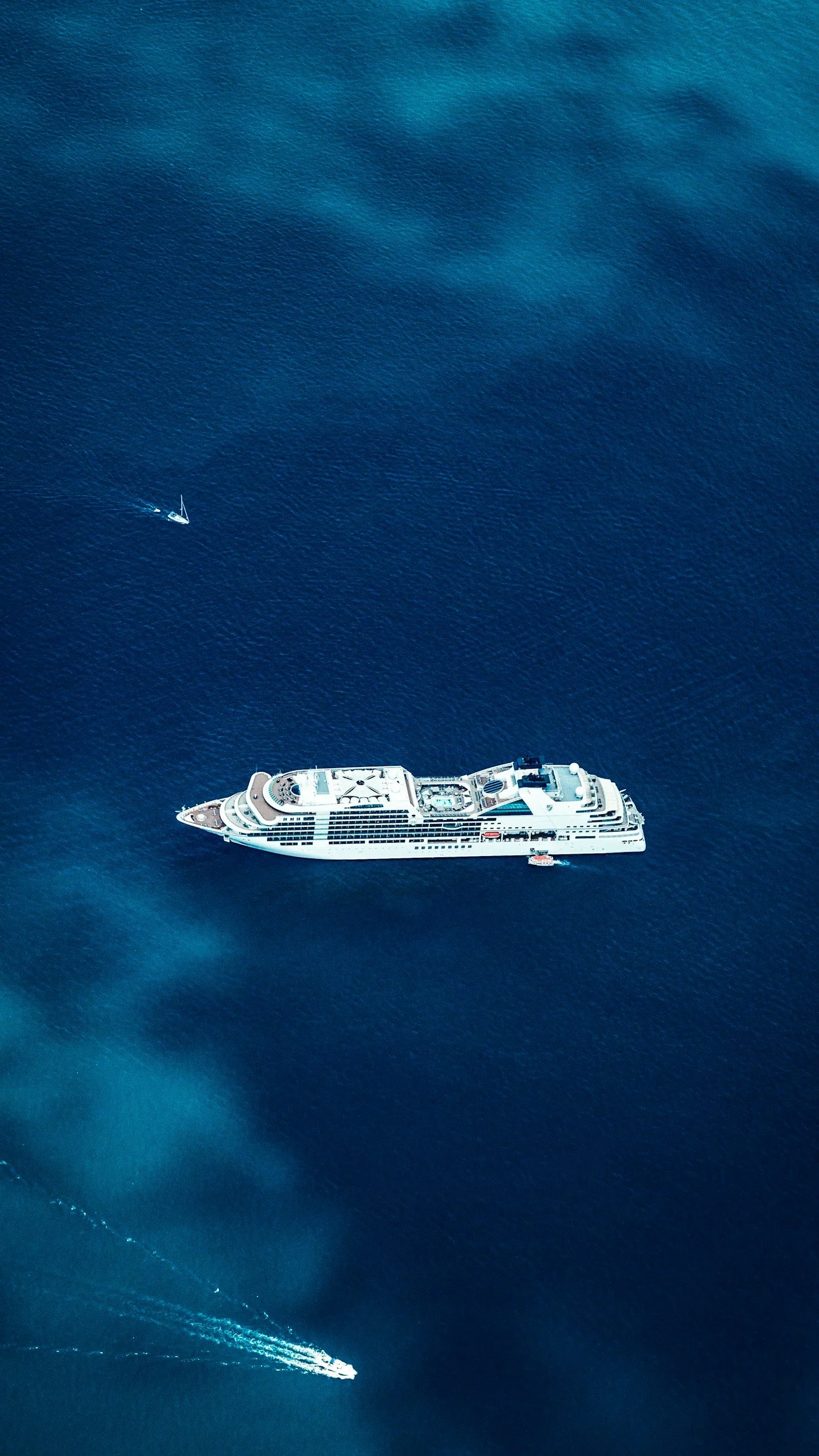 An aerial view of a large white cruise ship sailing in deep blue ocean water with a small boat nearby.