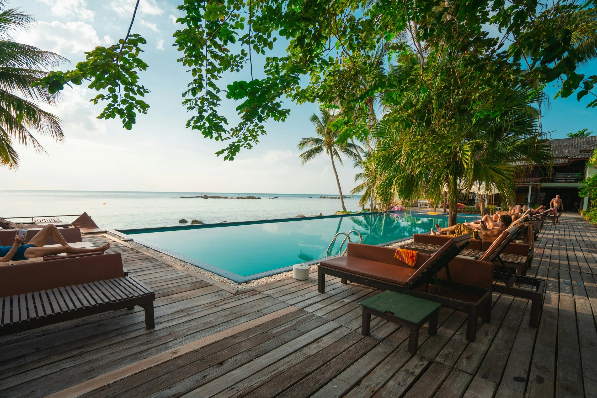 People relaxing on sun loungers around an outdoor infinity pool overlooking the ocean with palm trees and tropical foliage.