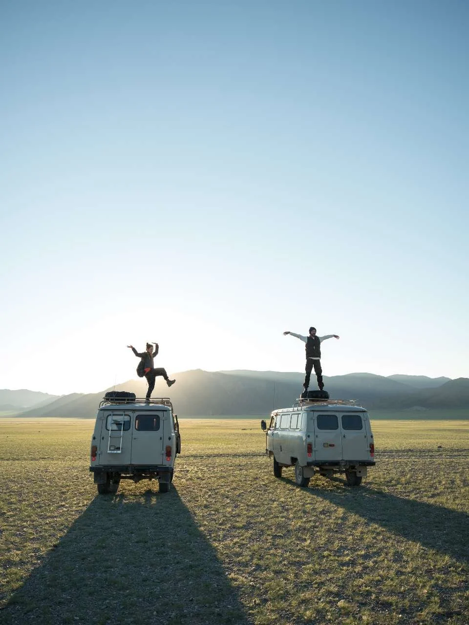 Two people standing on the roofs of two white vans in a vast open field with mountains in the distance during daytime.