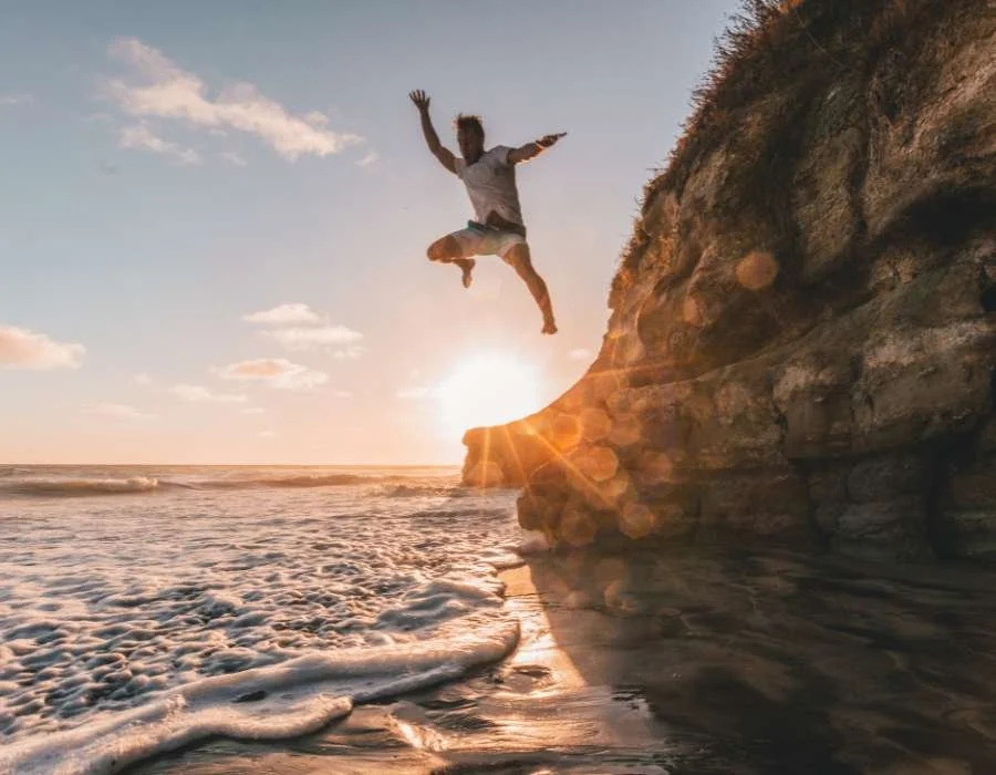 A person in mid-air jump on a beach at sunset near a rocky cliff.