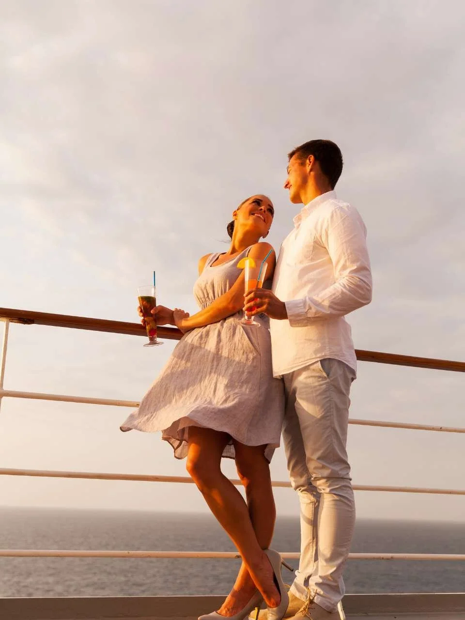 A man and woman on a balcony overlooking the ocean at sunset, holding drinks and smiling at each other.