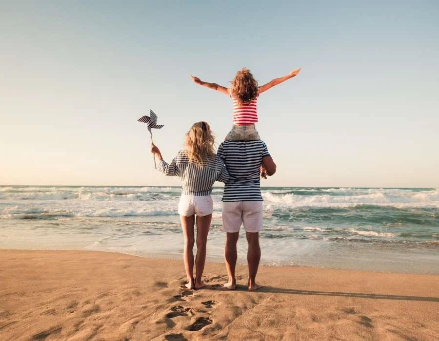 A family of three with two adults and a child at the beach during sunset, standing by the ocean shore and facing the water.