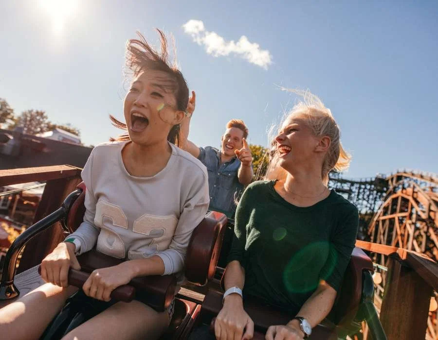 Three friends enjoying a roller coaster ride at an amusement park on a sunny day, smiling and laughing.