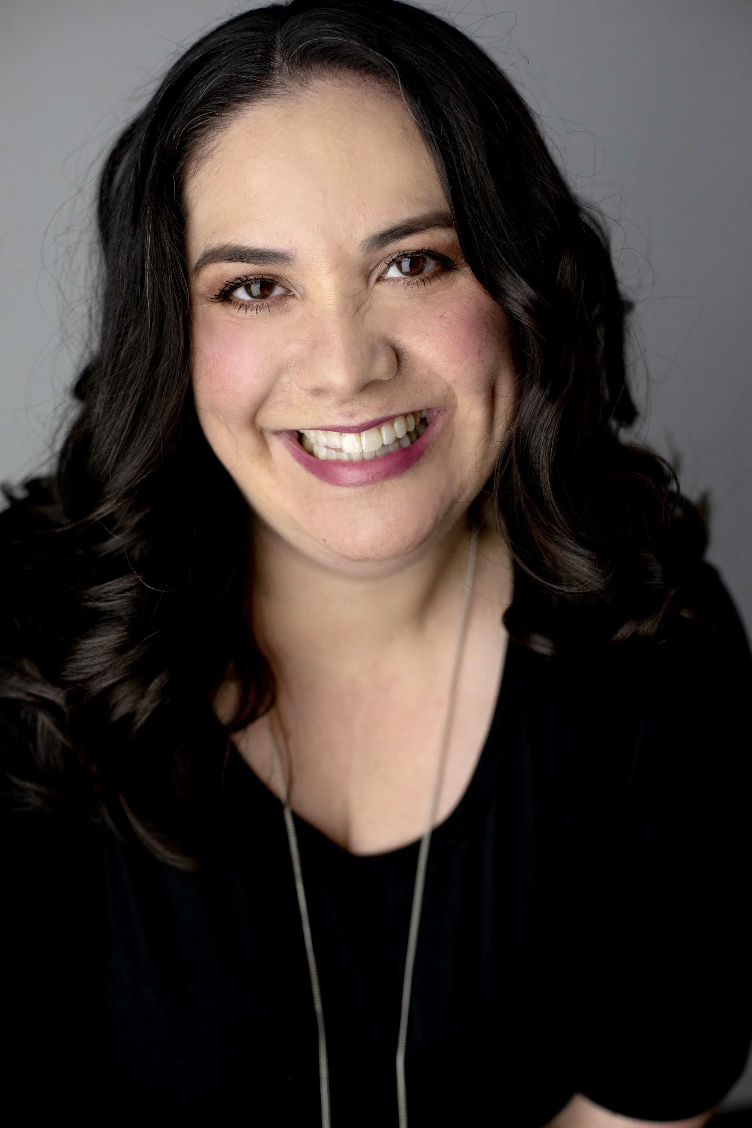 Sarah Sanders with wavy black hair, wearing a black top and a necklace, against a gray background.