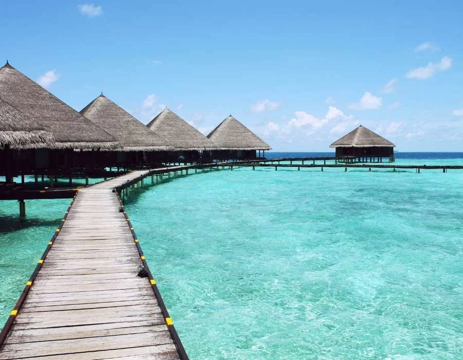 Overwater bungalows with thatched roofs connected by a wooden walkway over turquoise ocean water under a partly cloudy sky.