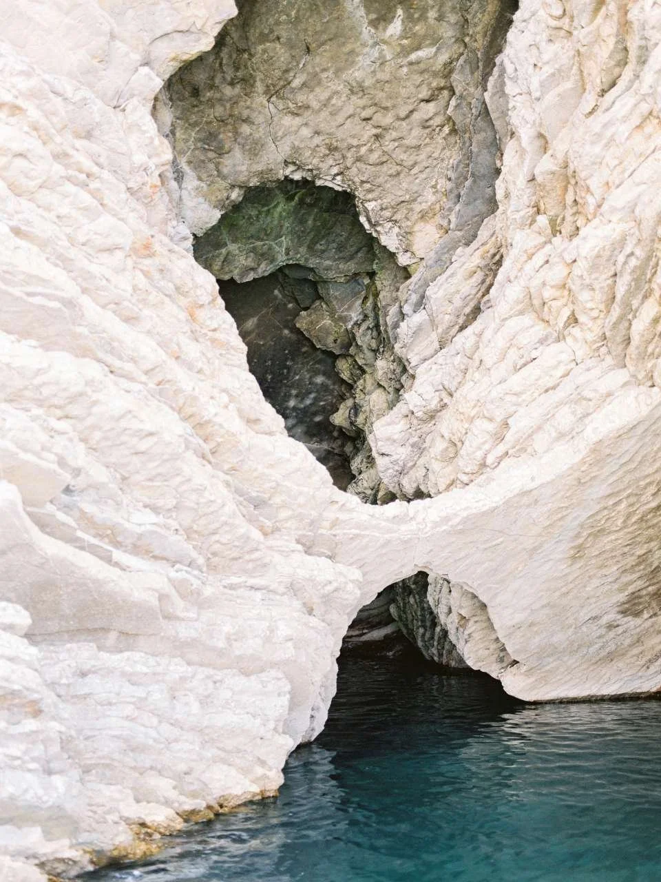 White rocky cliff with a natural arch leading to a small cave, with turquoise water at the base.