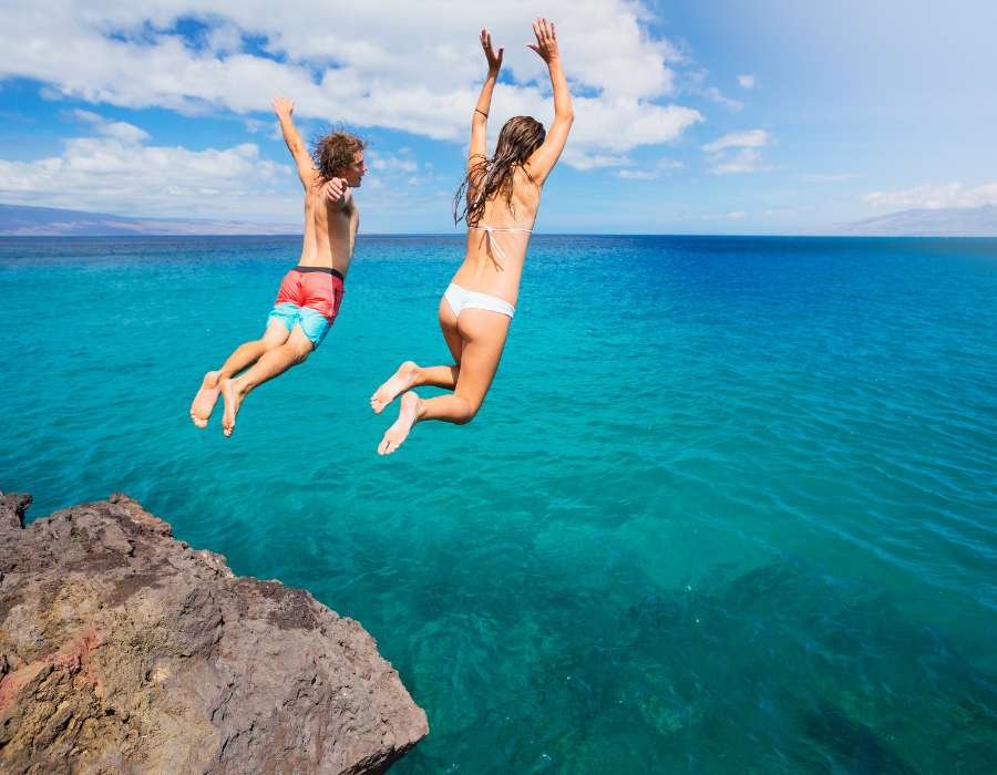 A man and woman jumping off a rocky cliff into the ocean on a bright, sunny day.