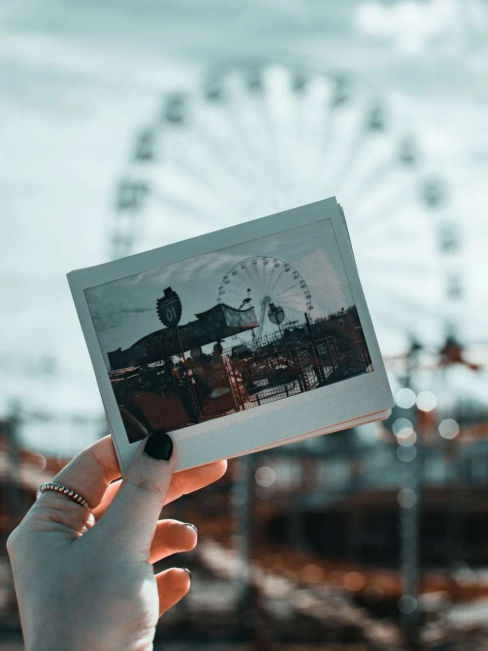 A person holding a polaroid photo of a carnival with a Ferris wheel, while the Ferris wheel is visible in the background.