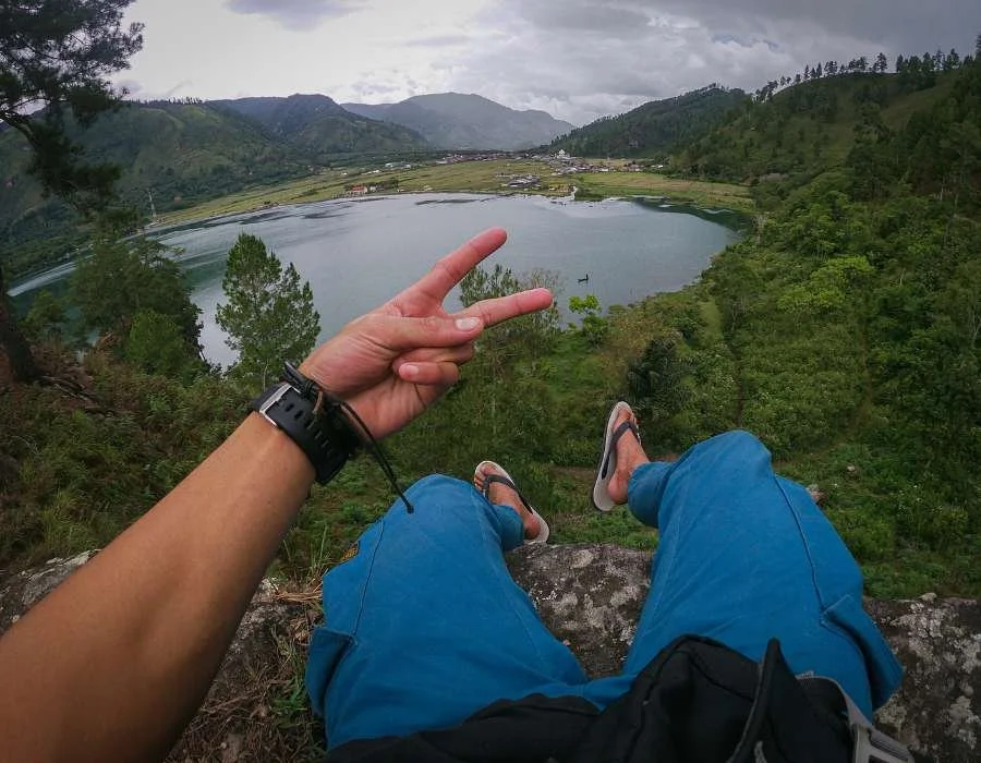 Person sitting on a ledge overlooking a lake surrounded by green hills and mountains, making a peace sign with their hand.