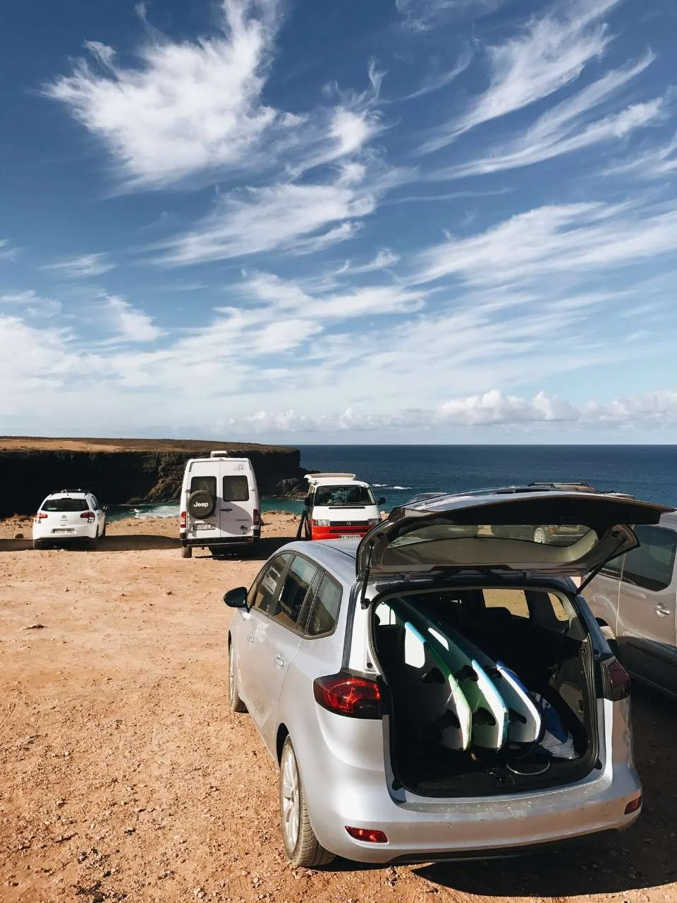 A silver hatchback car with a surfboard in the trunk, parked on a sandy area near the ocean. In the background, there are a few other vehicles and a rocky coastline with a partly cloudy sky overhead.