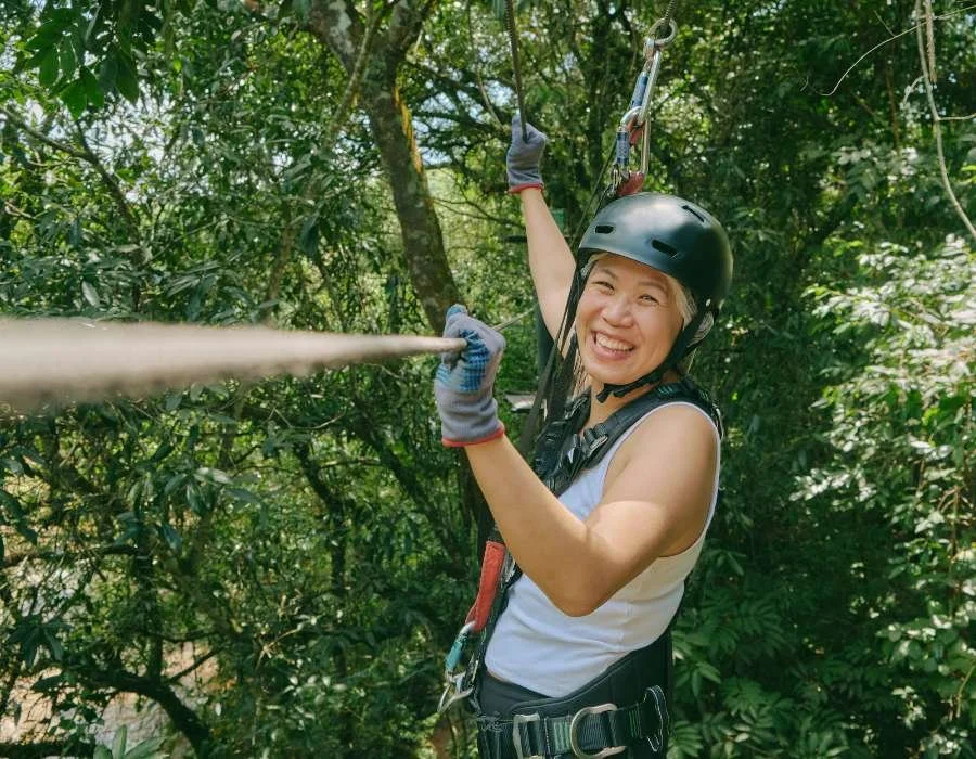 A woman wearing a helmet and gloves is zip lining through a green forest, smiling and holding onto the zip line cable.