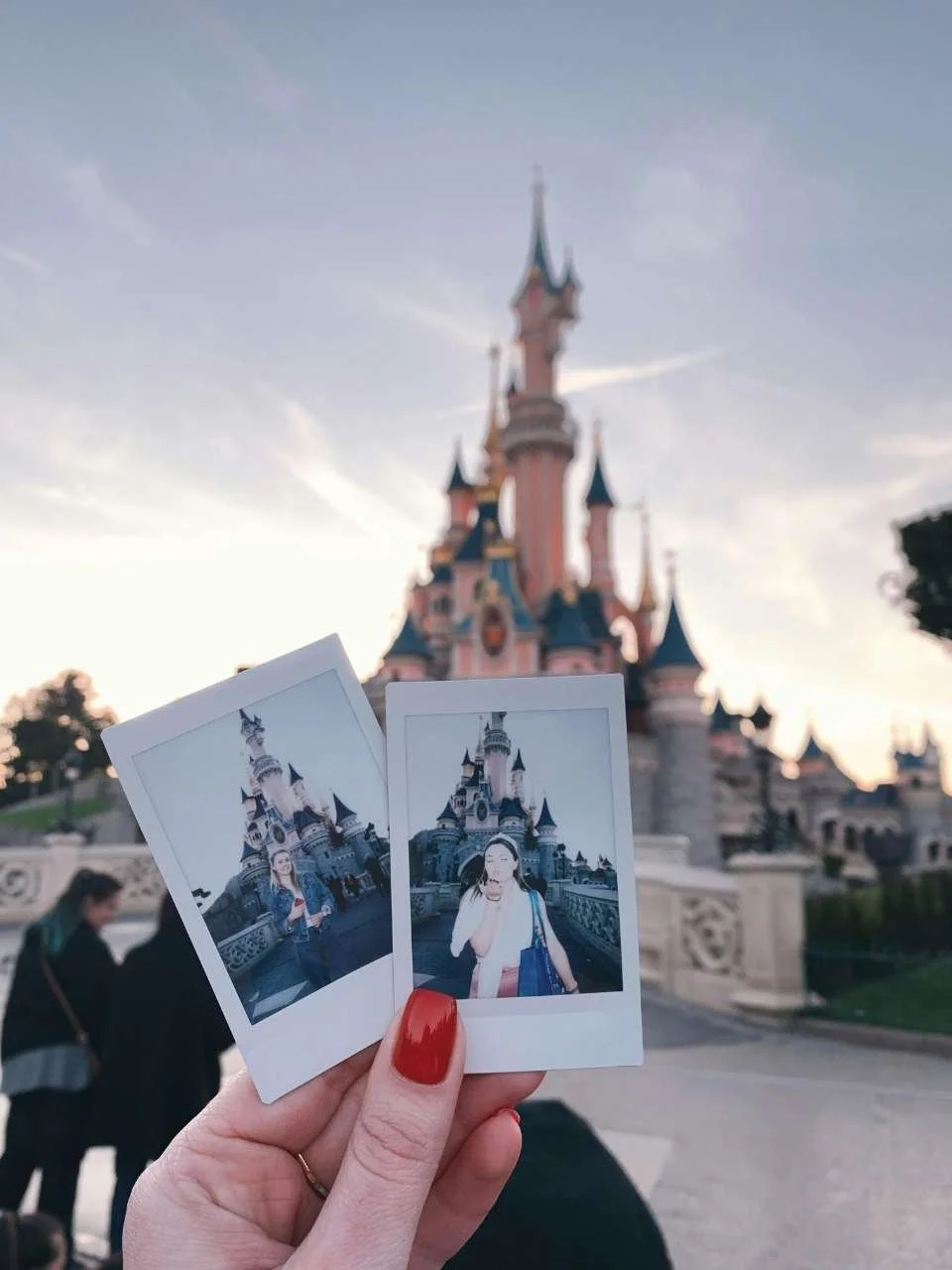 Person holding two instant photos in front of Sleeping Beauty Castle at Disneyland with other visitors in the background.