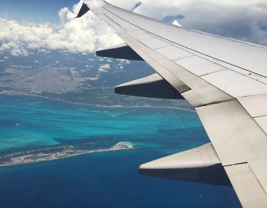 View of an airplane wing from the window showing turquoise water, a small island, and land below, with clouds in the sky.
