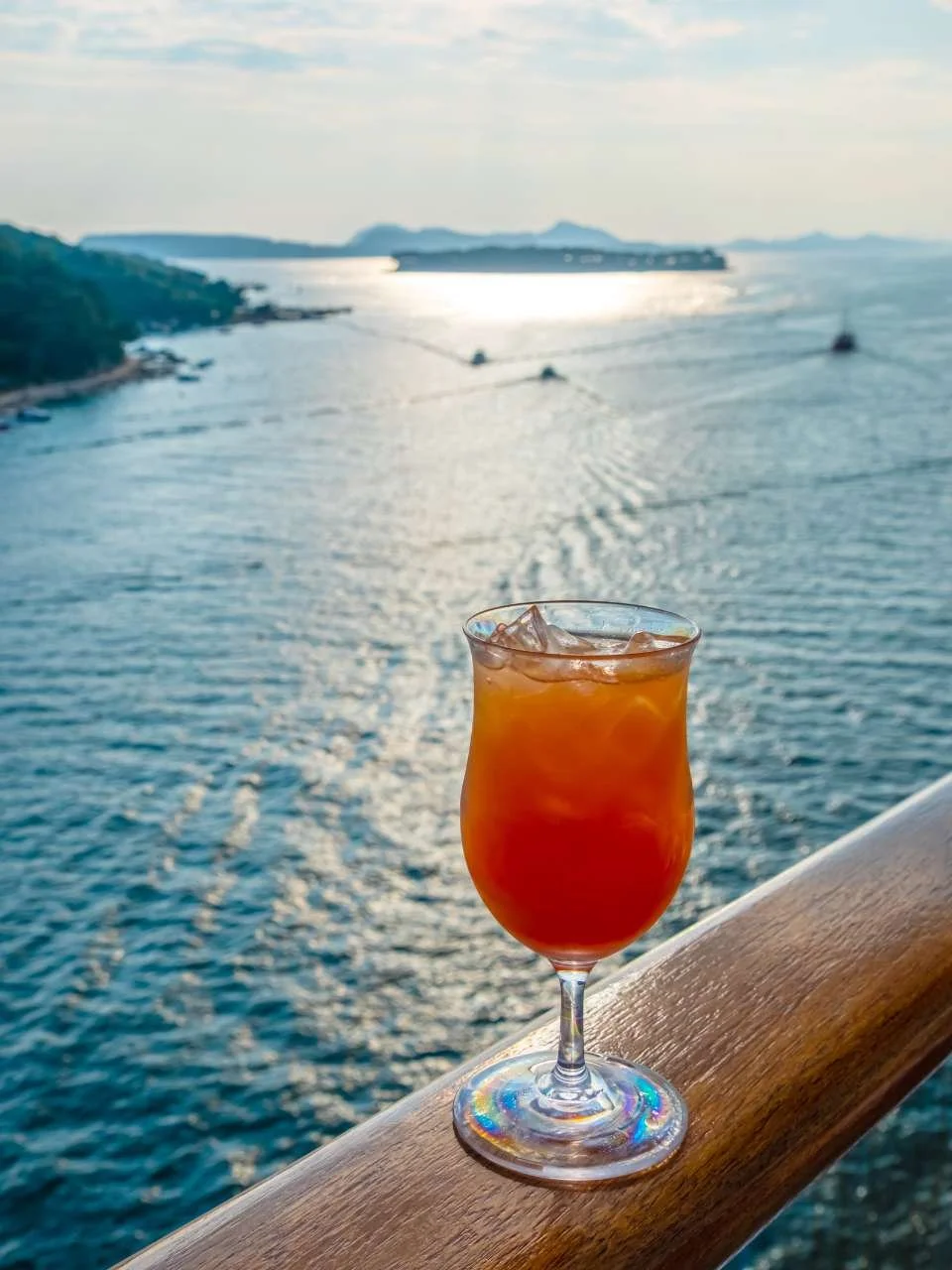 A cocktail glass filled with an orange-colored drink and ice, placed on a wooden railing overlooking a large body of water with boats and distant land forms, under a partly cloudy sky during sunset.