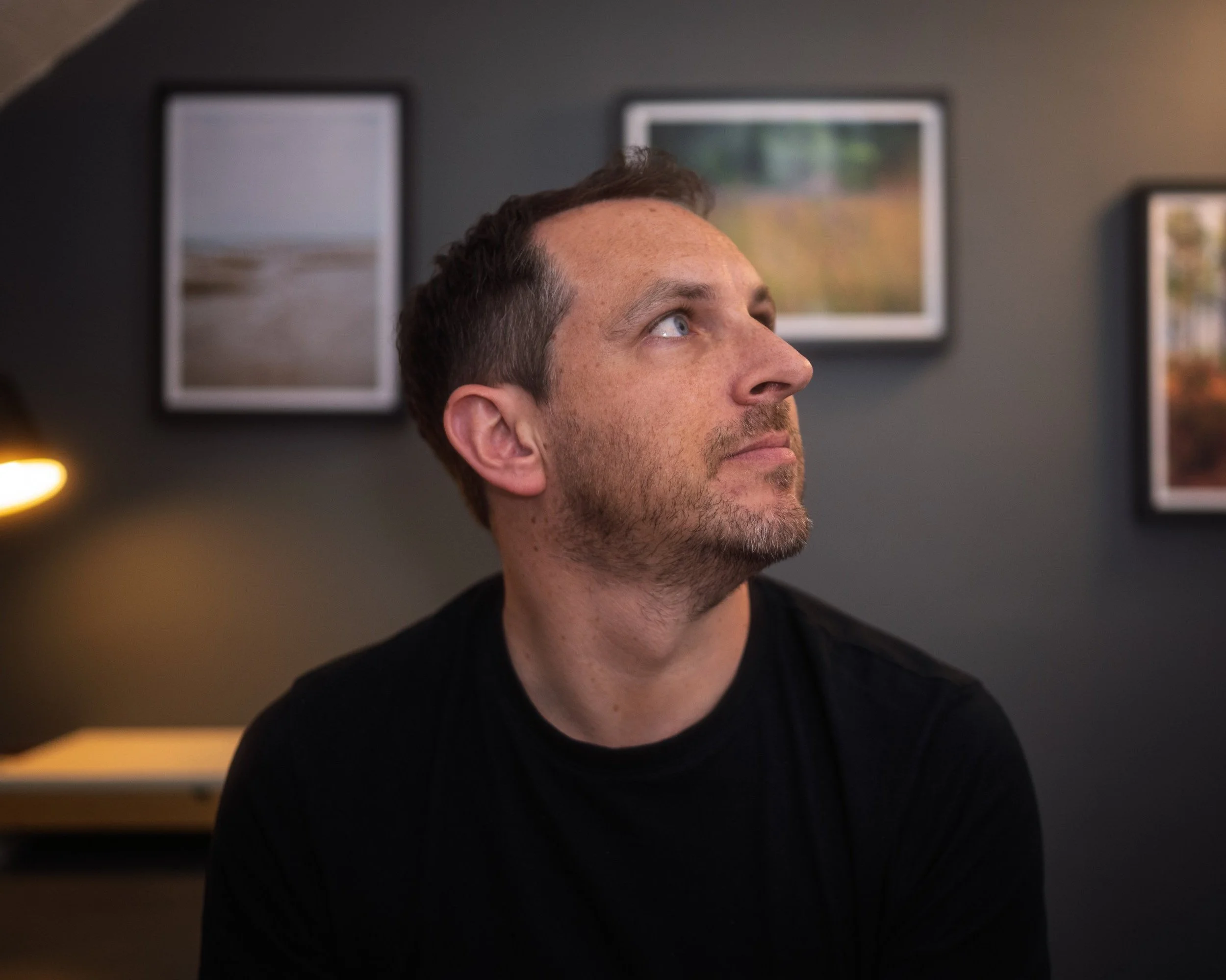 A man with short brown hair and a beard looking upwards, sitting in a room with dark walls and framed landscape pictures.
