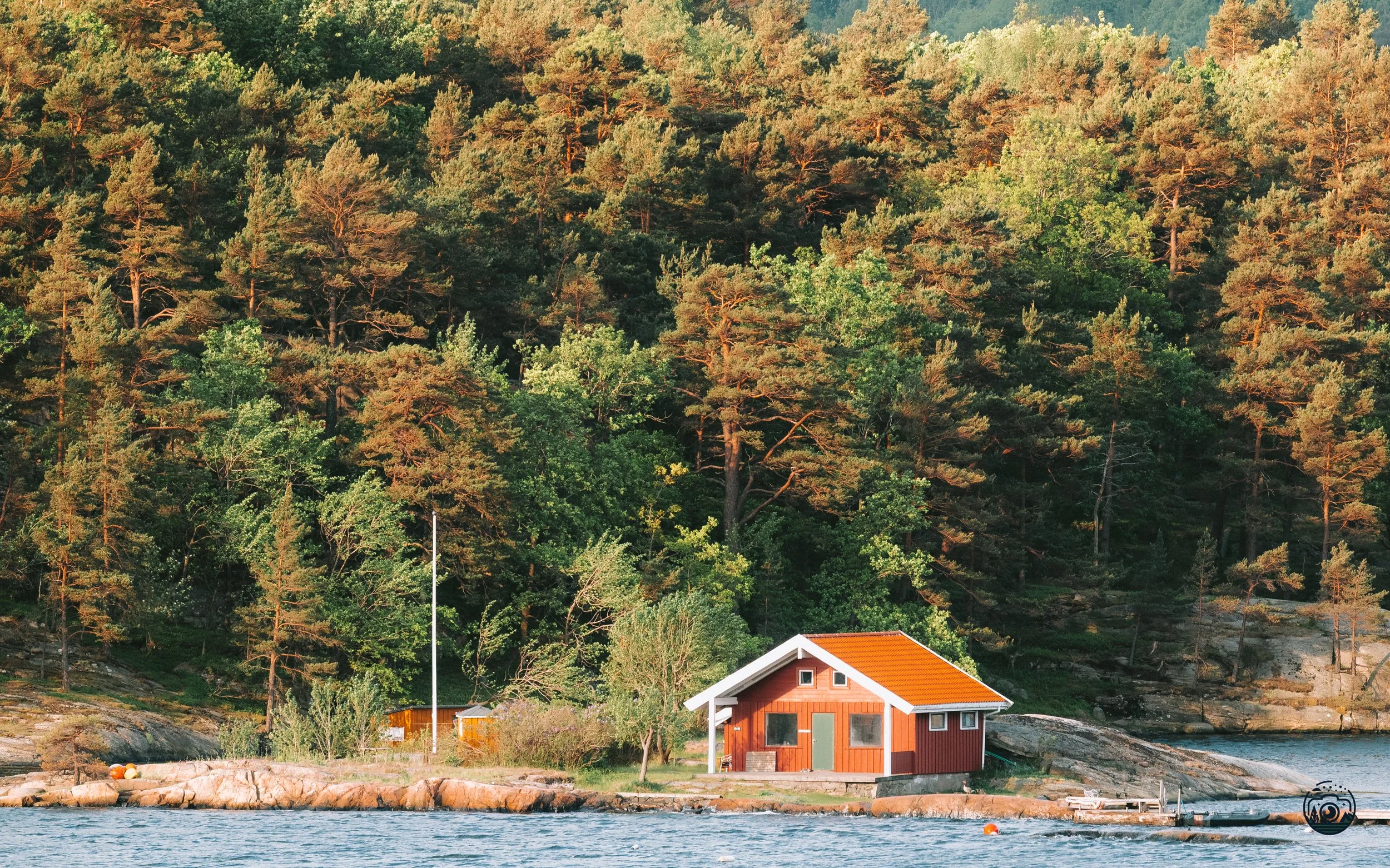 A red-roofed cabin tucked beside a calm shoreline, surrounded by tall pine trees catching the warm, golden-hour light.