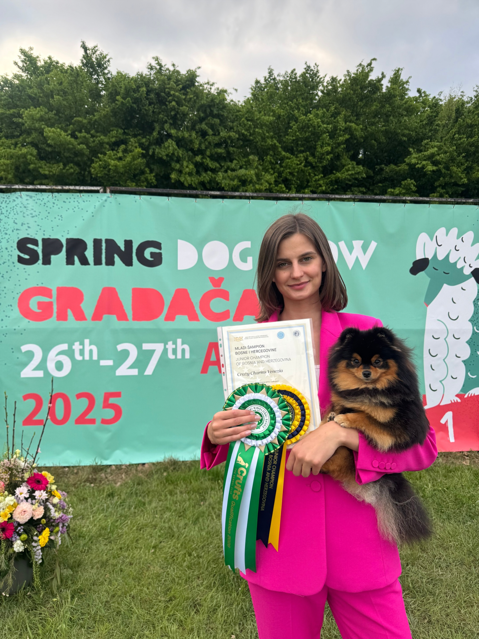 Une femme avec un chien de race Spitz allemand, tenant un certificat et des rubans d'obtention de prix, devant un panneau publicitaire pour un concours canin en Bosnie Herzegovine, avec un bouquet de fleurs à côté.