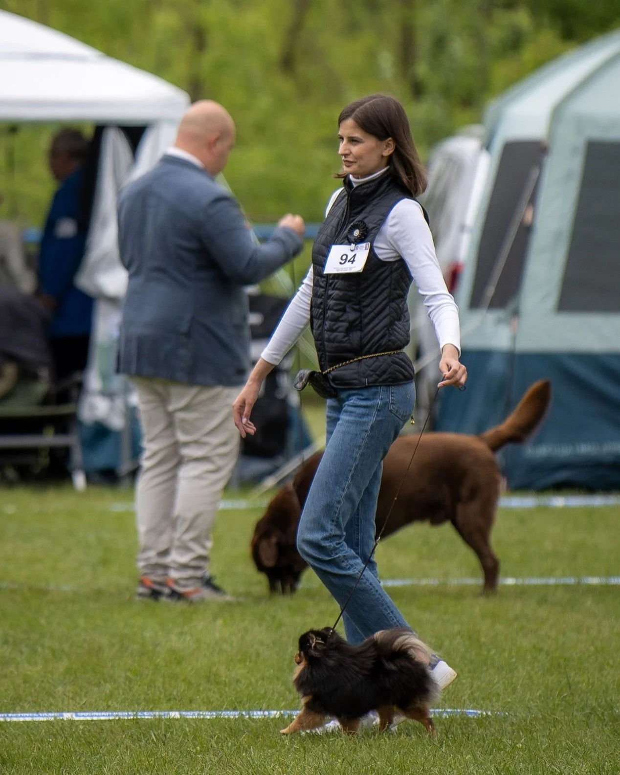 Une femme tenant une petite chienne spitz nain lors d'un concours canin en extérieur. Des tentes et des personnes en arrière-plan.