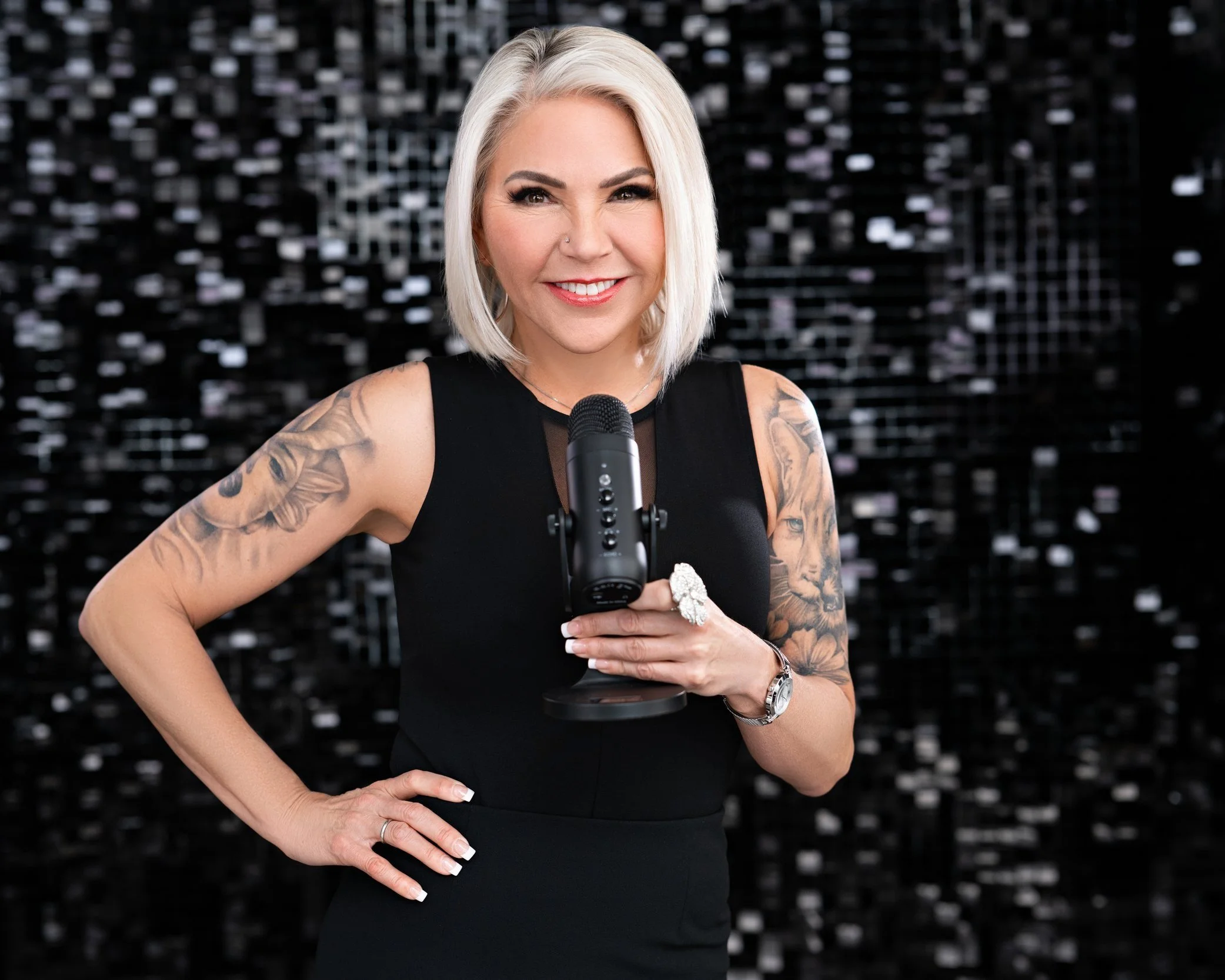 A smiling woman with blonde hair, tattoos on her arms, wearing a black dress, holding a microphone, standing in front of a dark, sequined background.