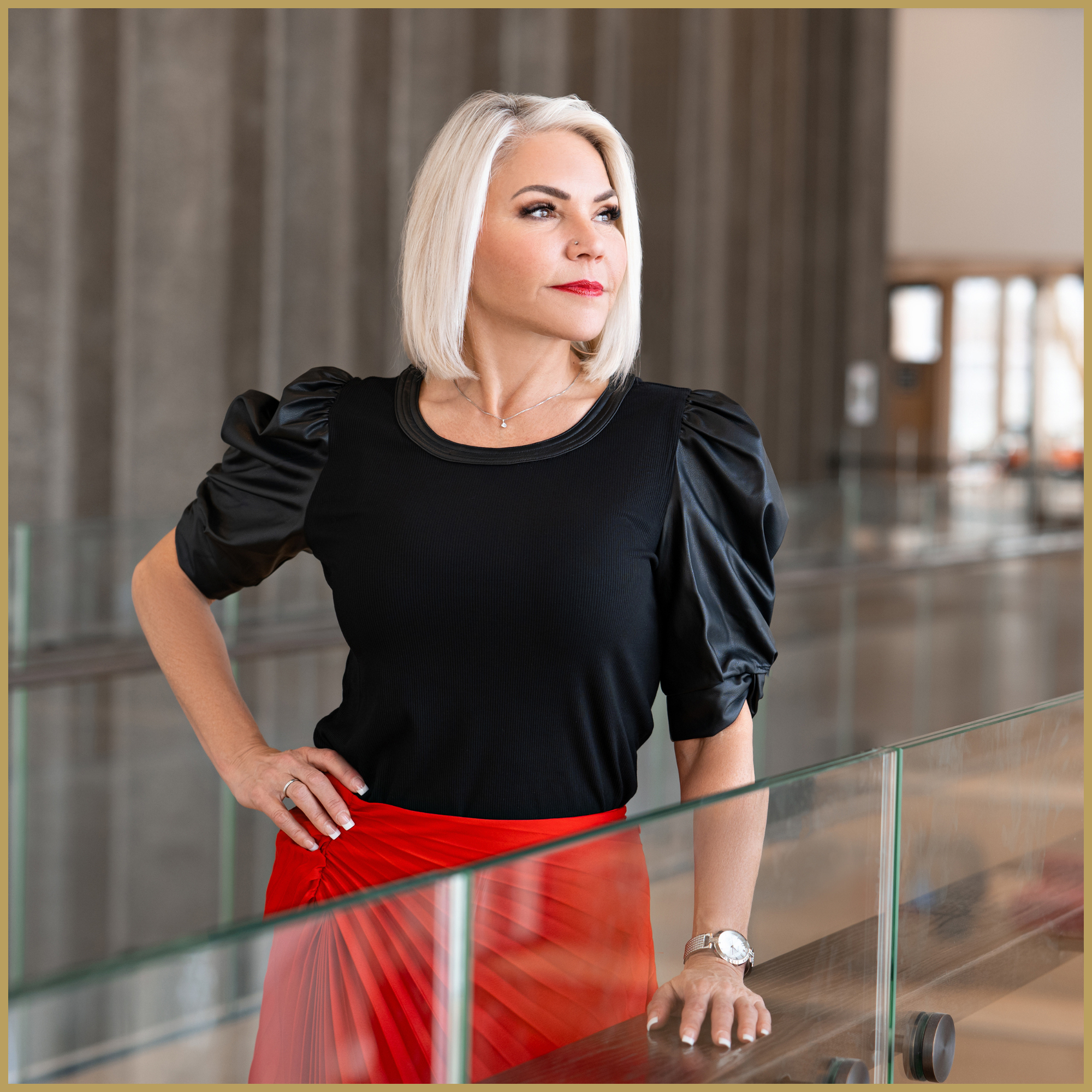 A mature woman with platinum blonde hair styled in a shoulder-length bob, wearing a black top with puffed sleeve details and a red pleated skirt, standing in a modern interior space with concrete walls and large windows. She is looking off to the side with a confident expression, resting her hand on a glass railing.