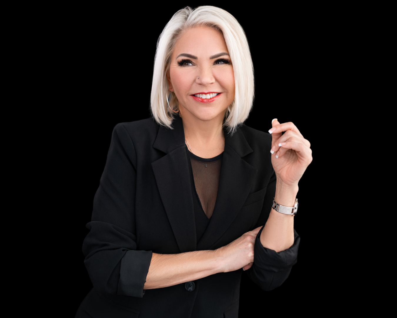 A smiling blonde woman with shoulder-length hair, wearing a black blazer and a black top with a sheer neckline, stands against a black background.