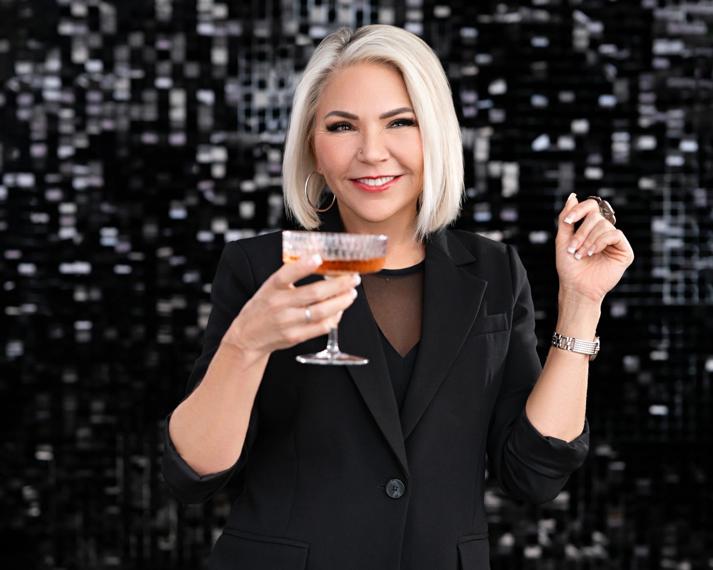 A woman with blonde hair holding a cocktail glass, smiling, dressed in black, standing in front of a dark, reflective background.